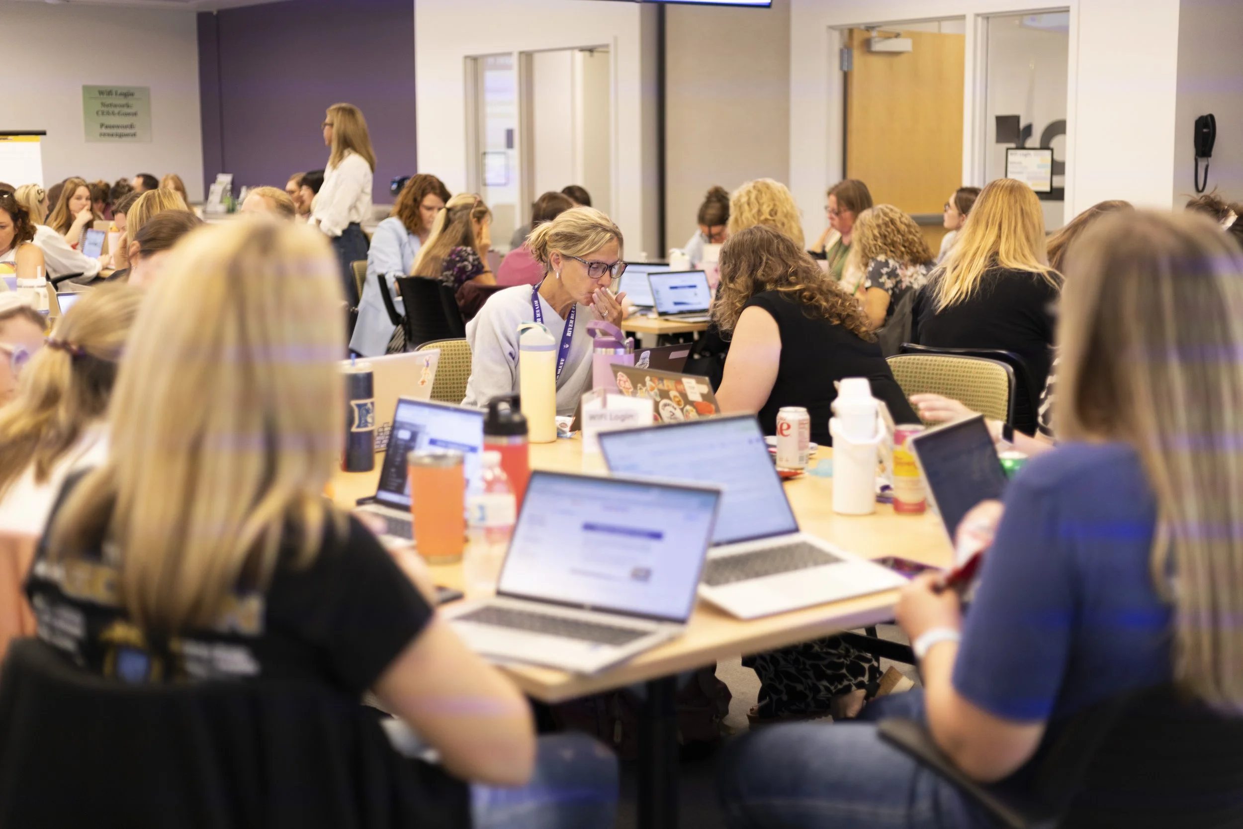 A large group of women are gathered around tables in a conference room, working on laptops and engaged in discussion. Some women are focused on their screens, while others are talking or taking notes.