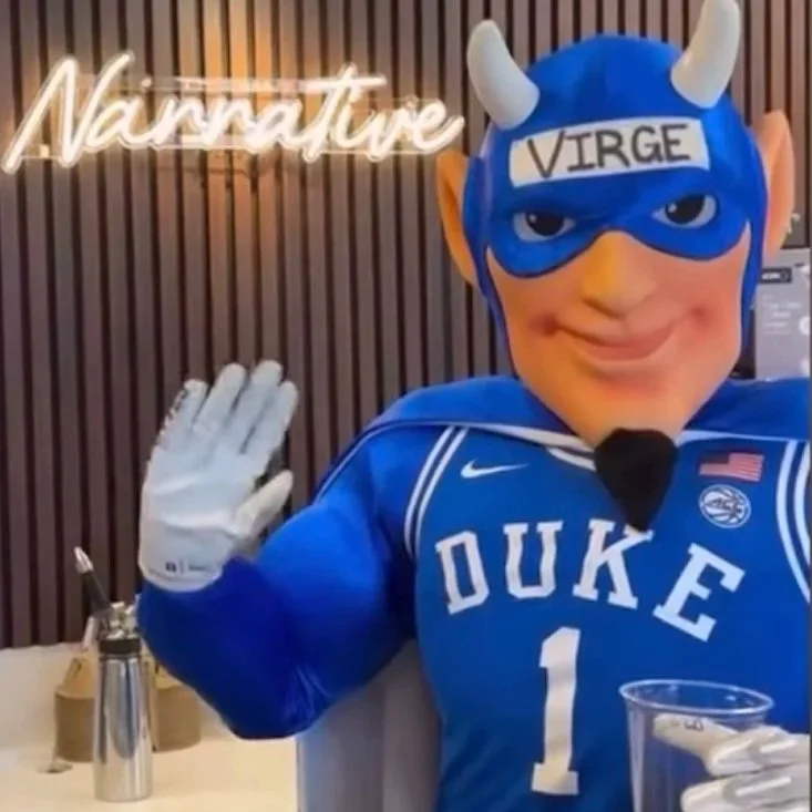 A mascot dressed in a Duke University sports jersey, wearing a blue superhero mask labeled "VIRGE" with small white horns, waving with a smile in a restaurant or cafe setting.