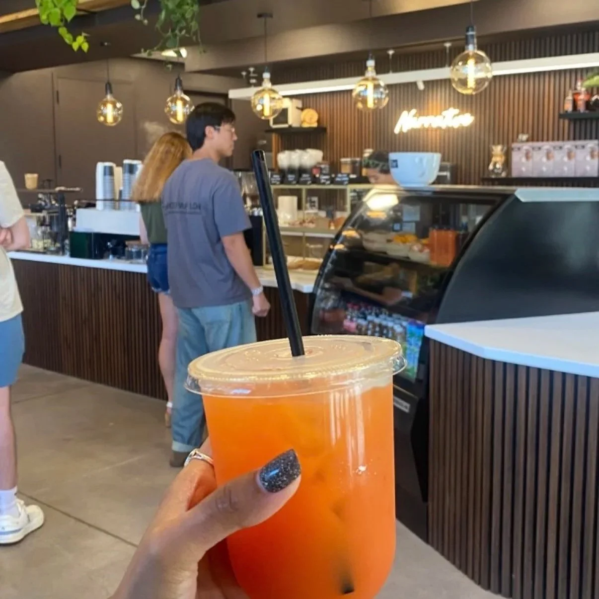 Point of view of a person holding an orange-colored drink with a black straw in a coffee shop. Behind the counter, there are customers and a barista preparing orders. The shop has a modern design with wooden accents and hanging pendant lights.
