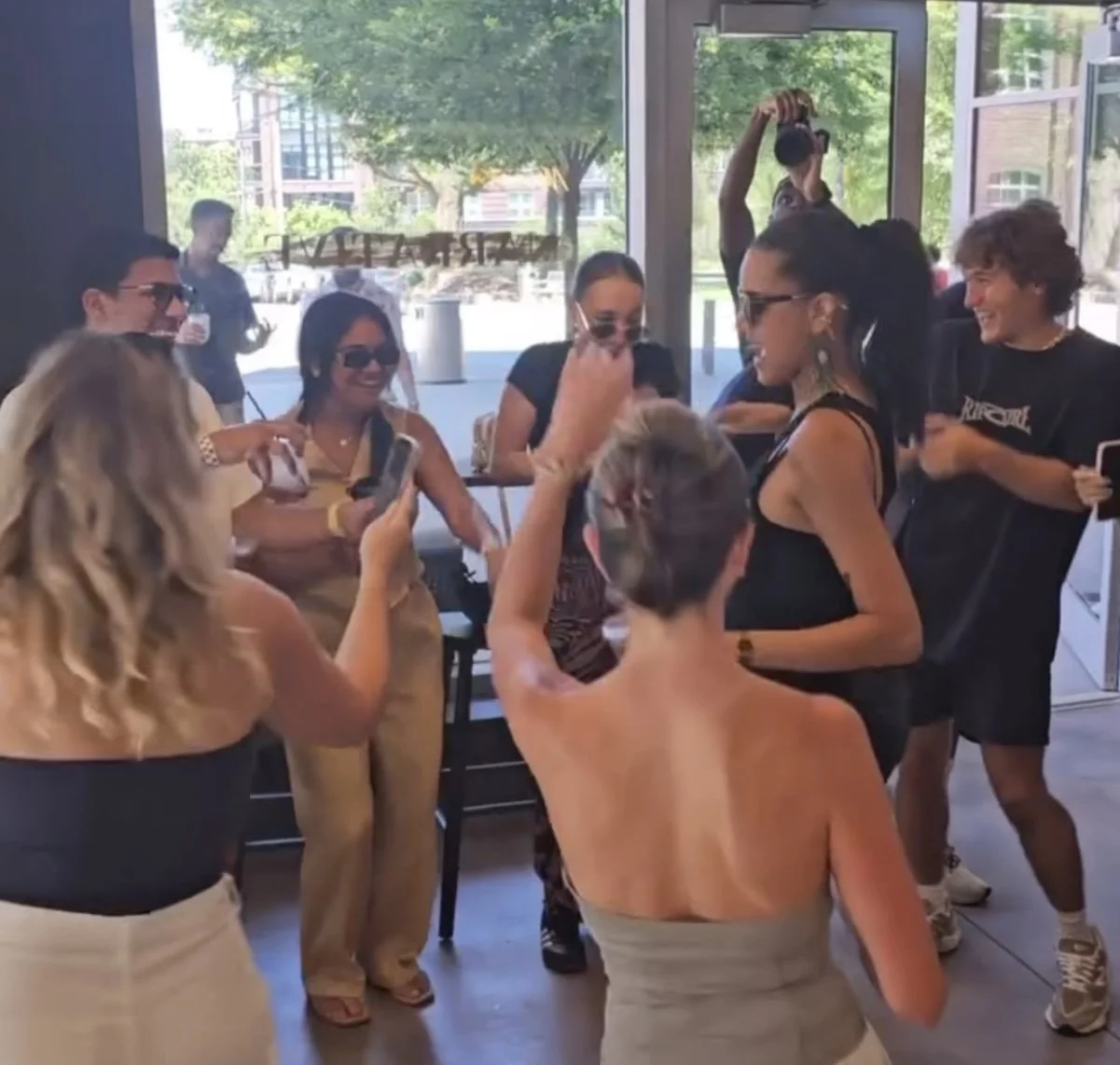 A group of people gathered inside a store or cafe, some holding phones, with large front windows showing trees and buildings outside, and a woman in a black dress with glasses standing and smiling.