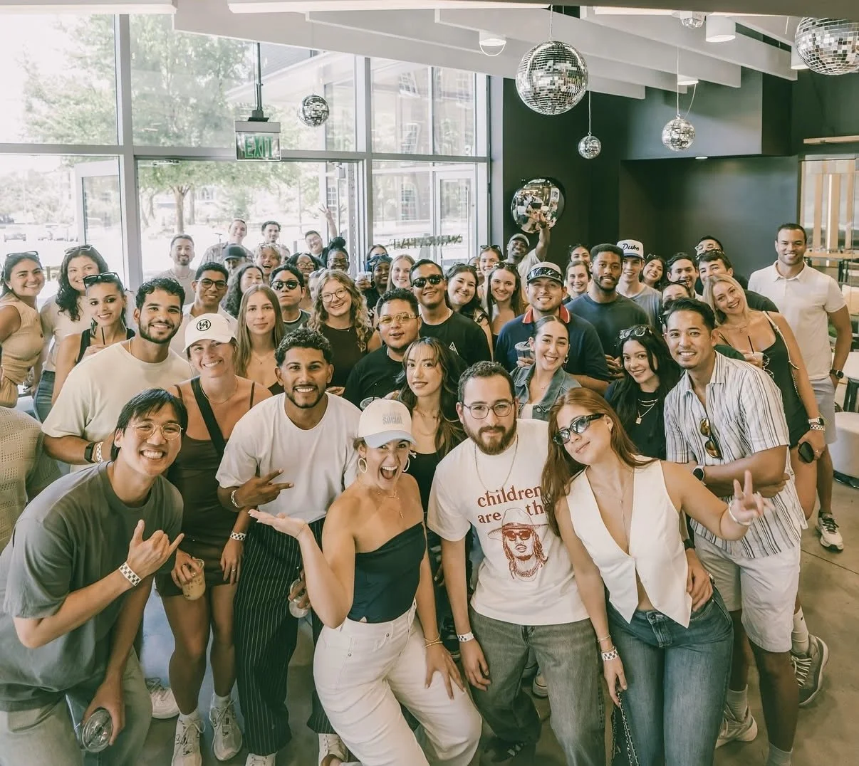 A large group of smiling people posing indoors near large windows with natural light, some wearing sunglasses and casual summer clothing, in a festive atmosphere with disco balls hanging from the ceiling.