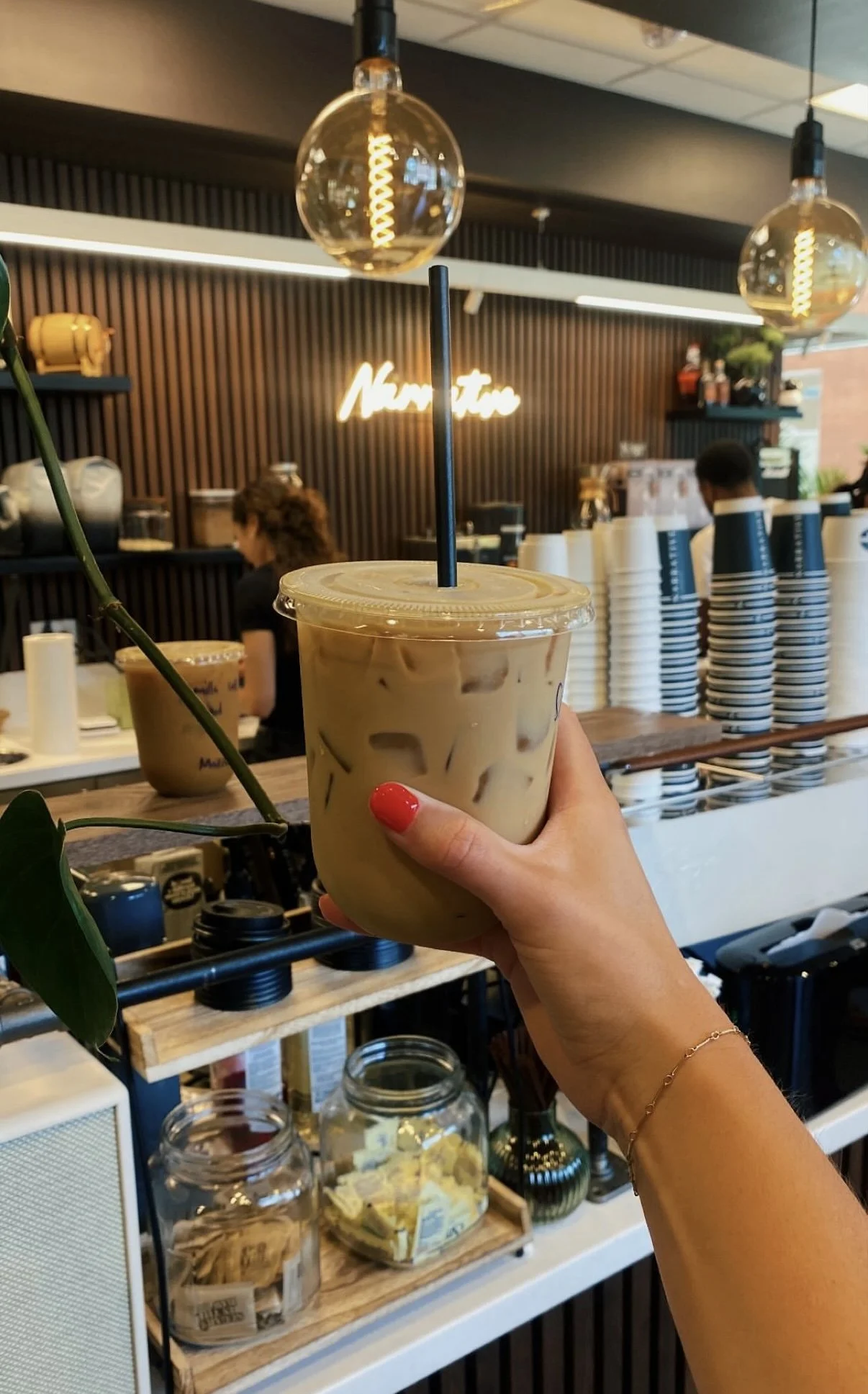 A person holding a plastic cup of iced coffee with milk and ice in a cafe. The cafe has a modern interior with hanging light bulbs and a wooden counter.