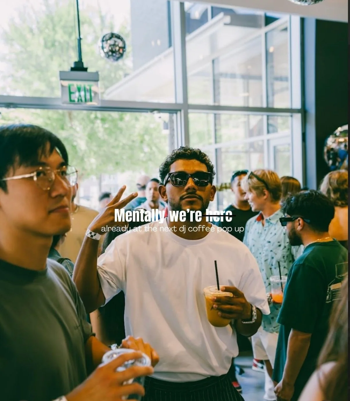 People gathered at a coffee shop, one man with sunglasses holding a drink, others engaged in conversations, with large windows and an "EXIT" sign visible inside the cafe.