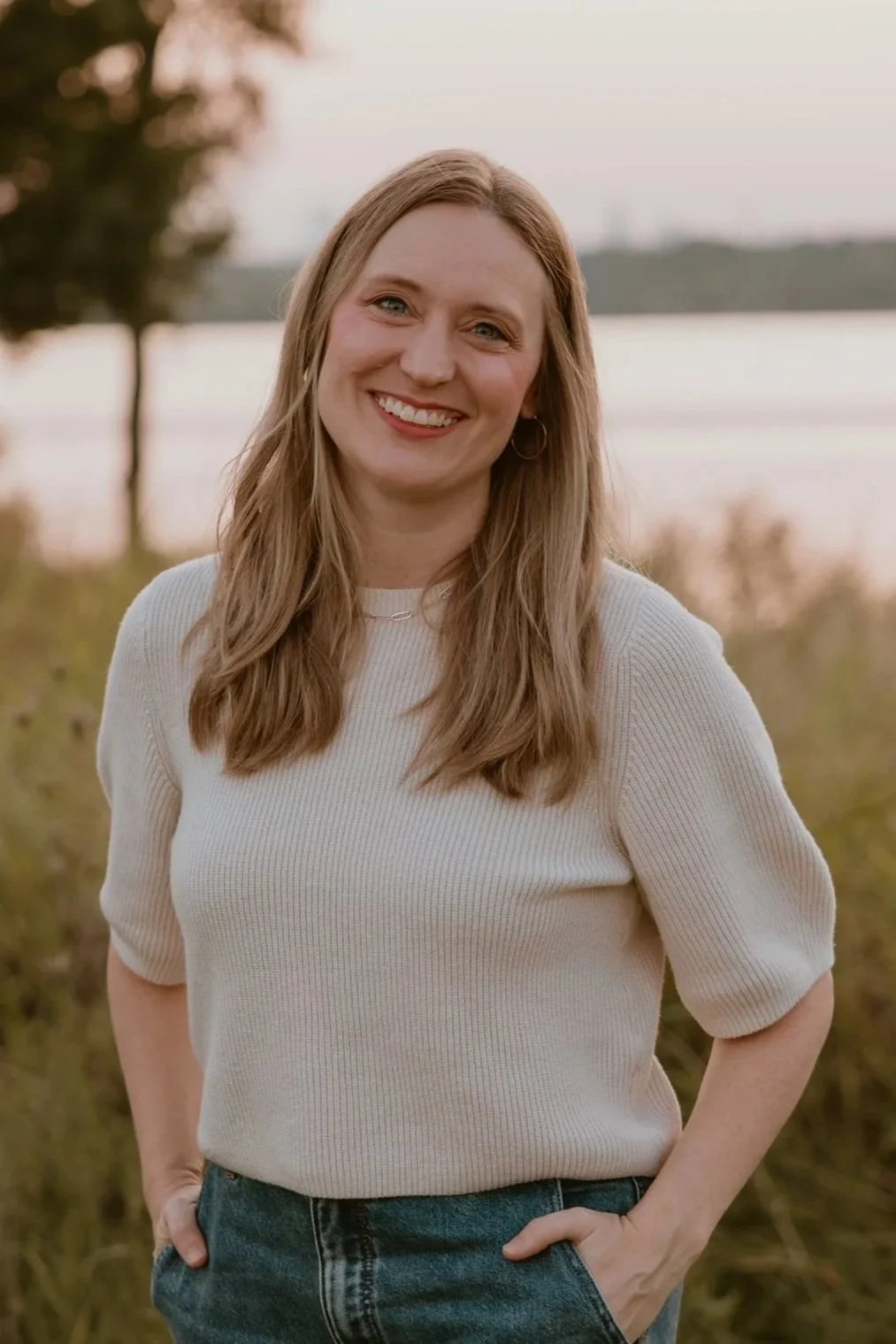 A young woman with long blonde hair smiling outdoors near a body of water at sunset, wearing a cream-colored sweater and blue jeans.