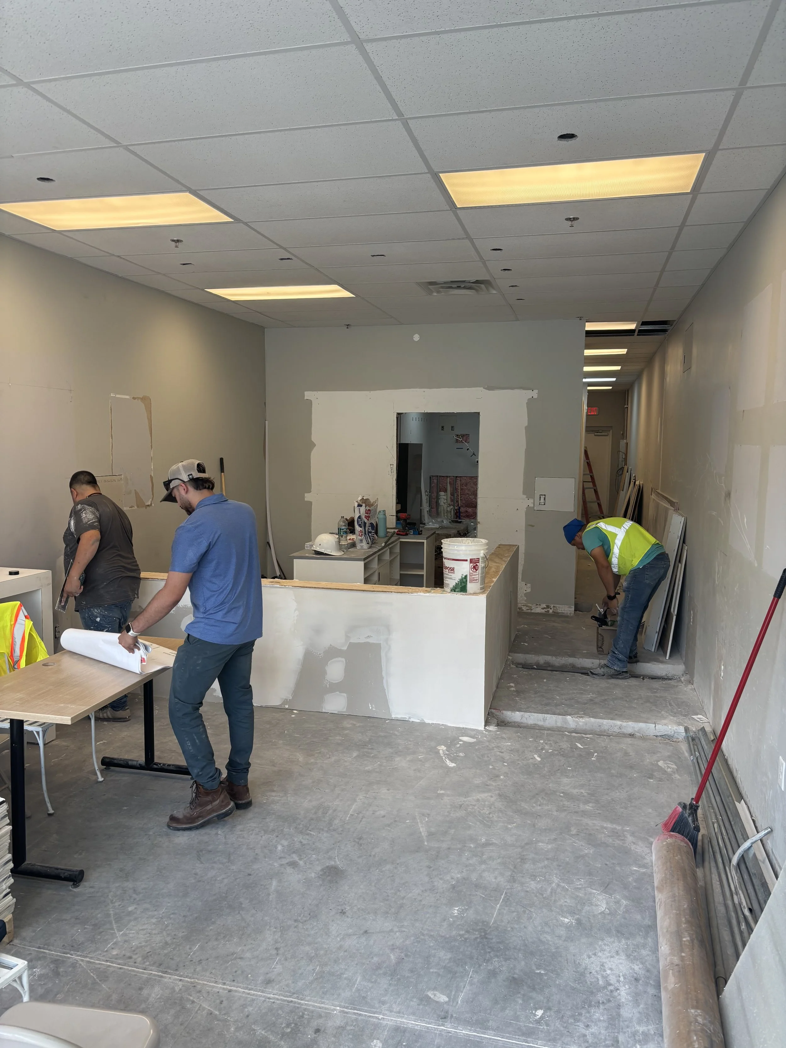 Interior of a construction site with workers installing drywall and flooring, tools and materials scattered, under fluorescent lighting.