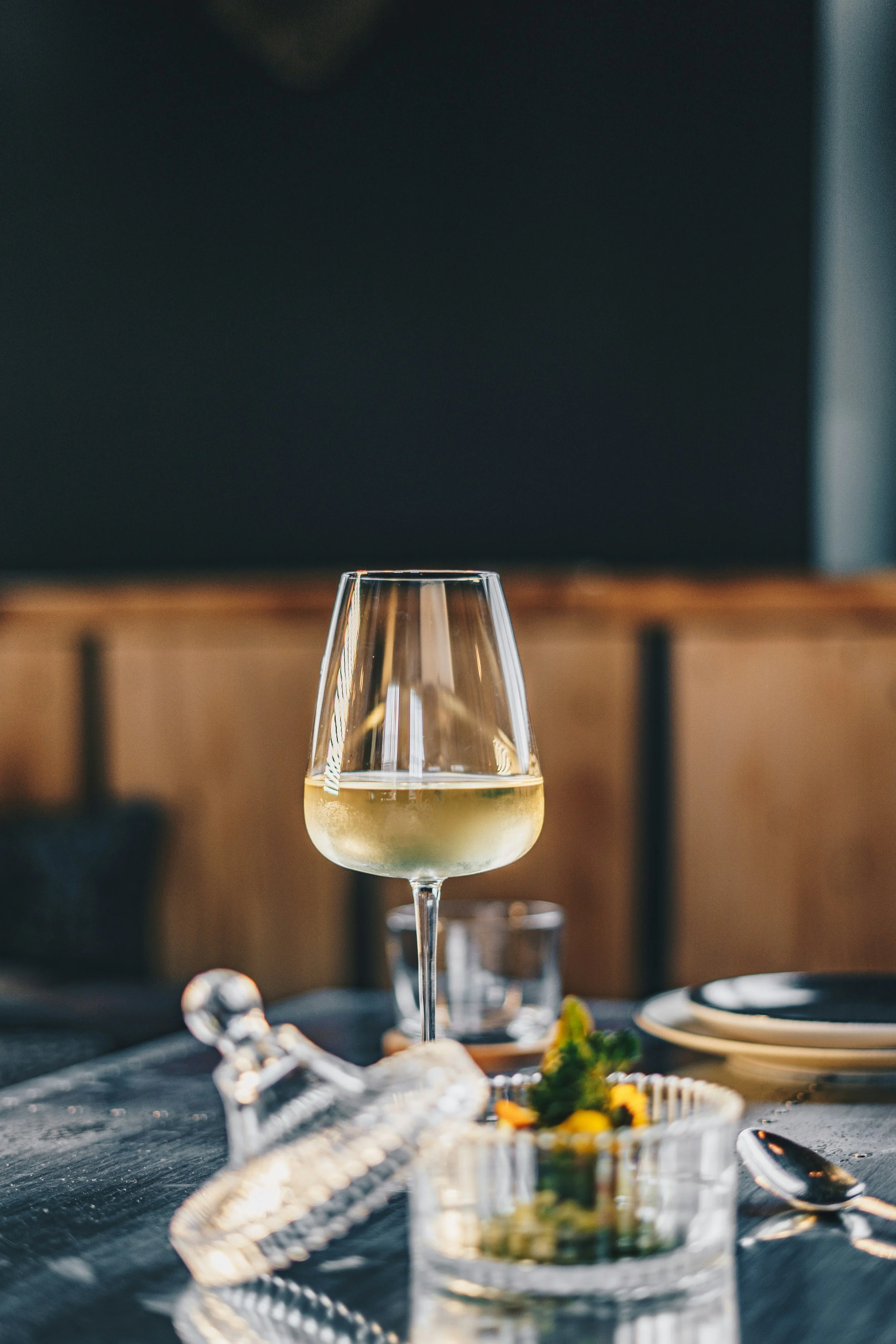 Close-up of a wine glass with white wine on a restaurant table, accompanied by a small dish and a spoon.