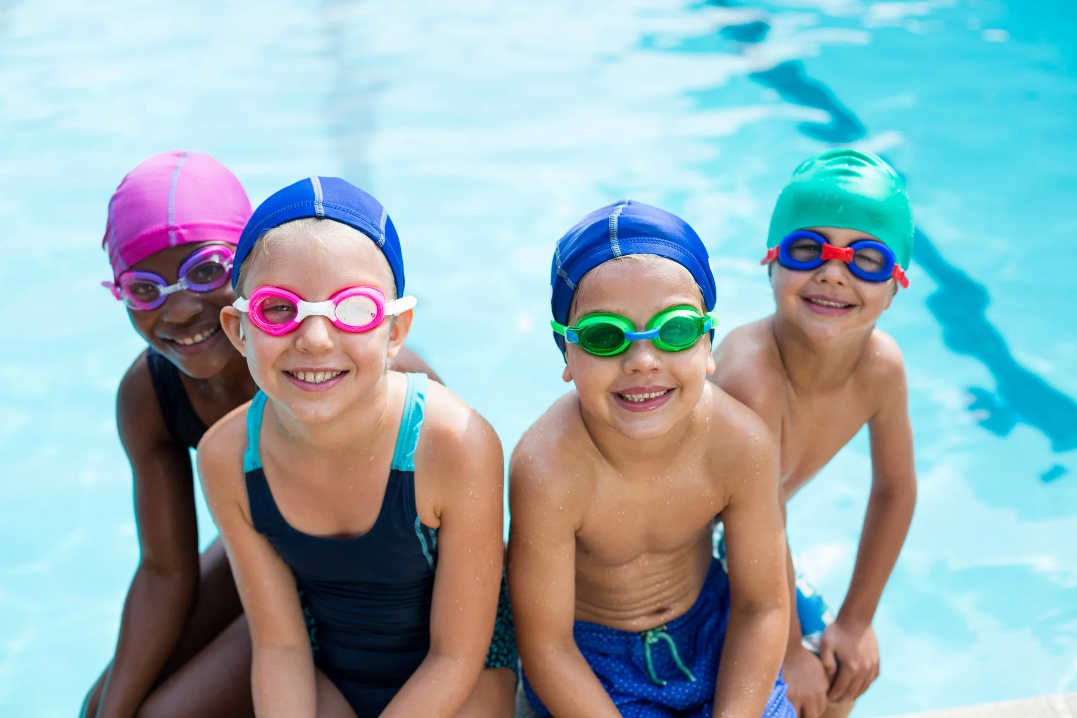 Four children in colorful swim caps and goggles smiling at the poolside.