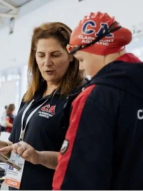 A woman and a young swimmer in a red swim cap and USA swimming jacket look at a card or paper together.