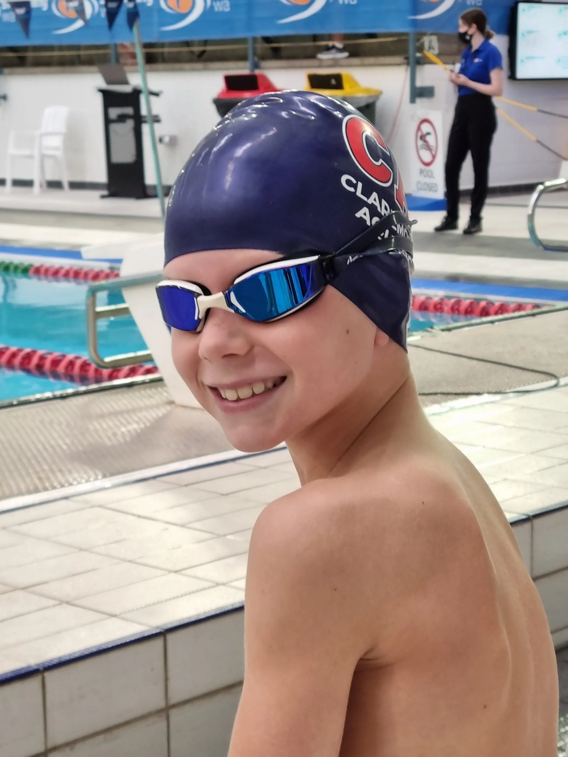 A young swimmer wearing a blue swim cap and blue goggles smiling at the poolside.