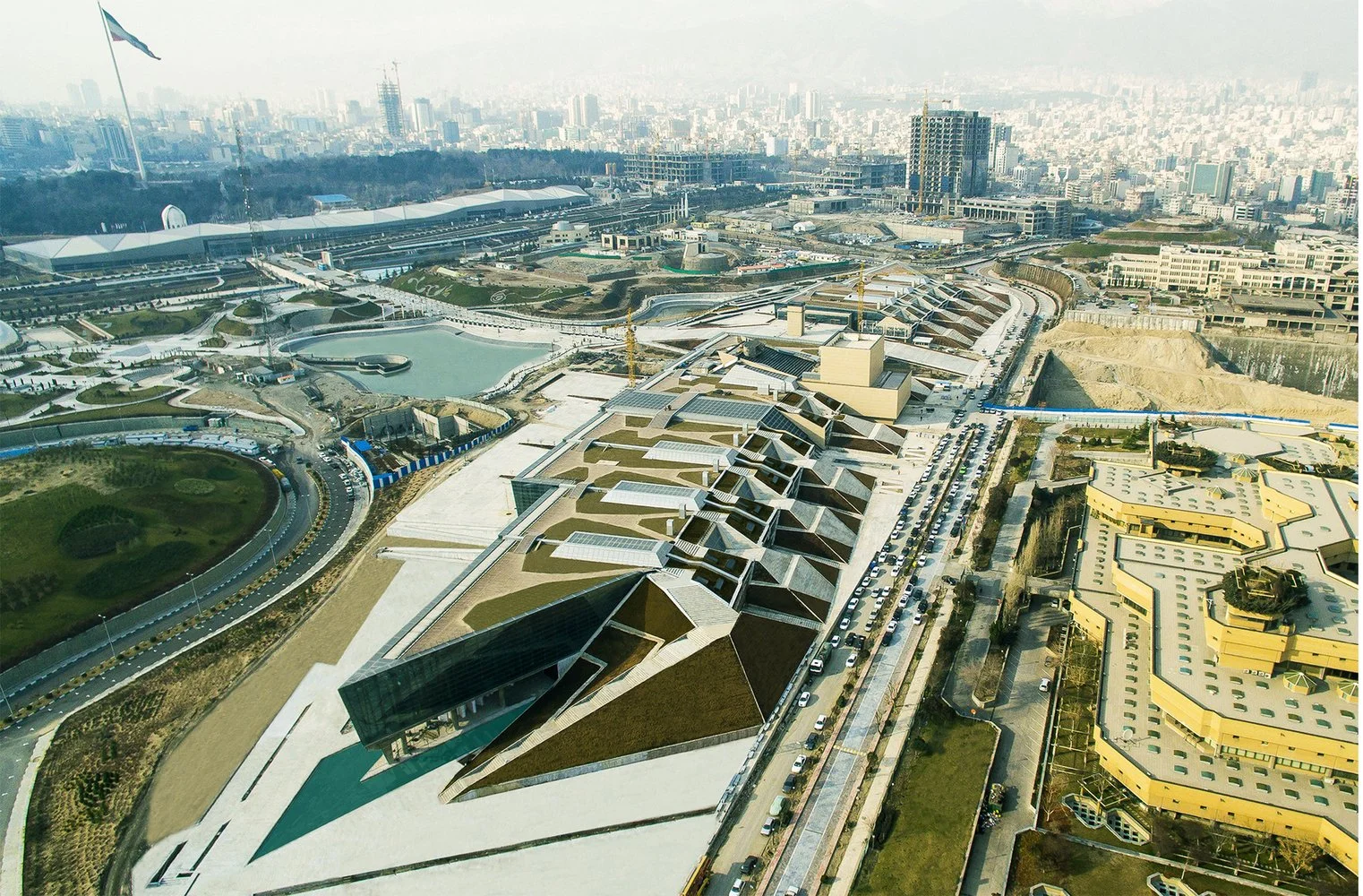 Aerial view of modern architectural complex in a city setting with roadways and an urban skyline in the background.