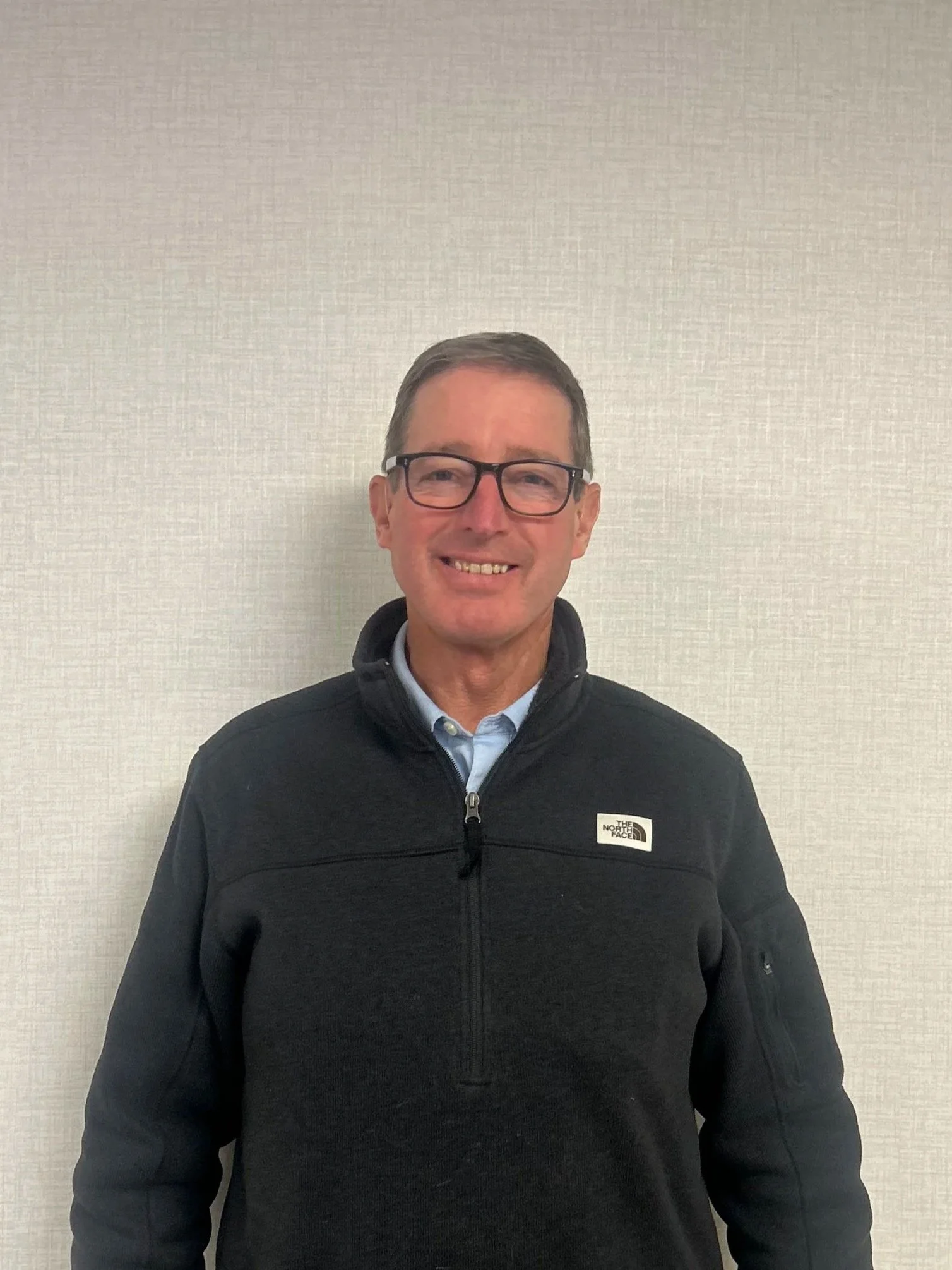 A smiling man with glasses wearing a black North Face jacket standing against a plain light-colored wall.