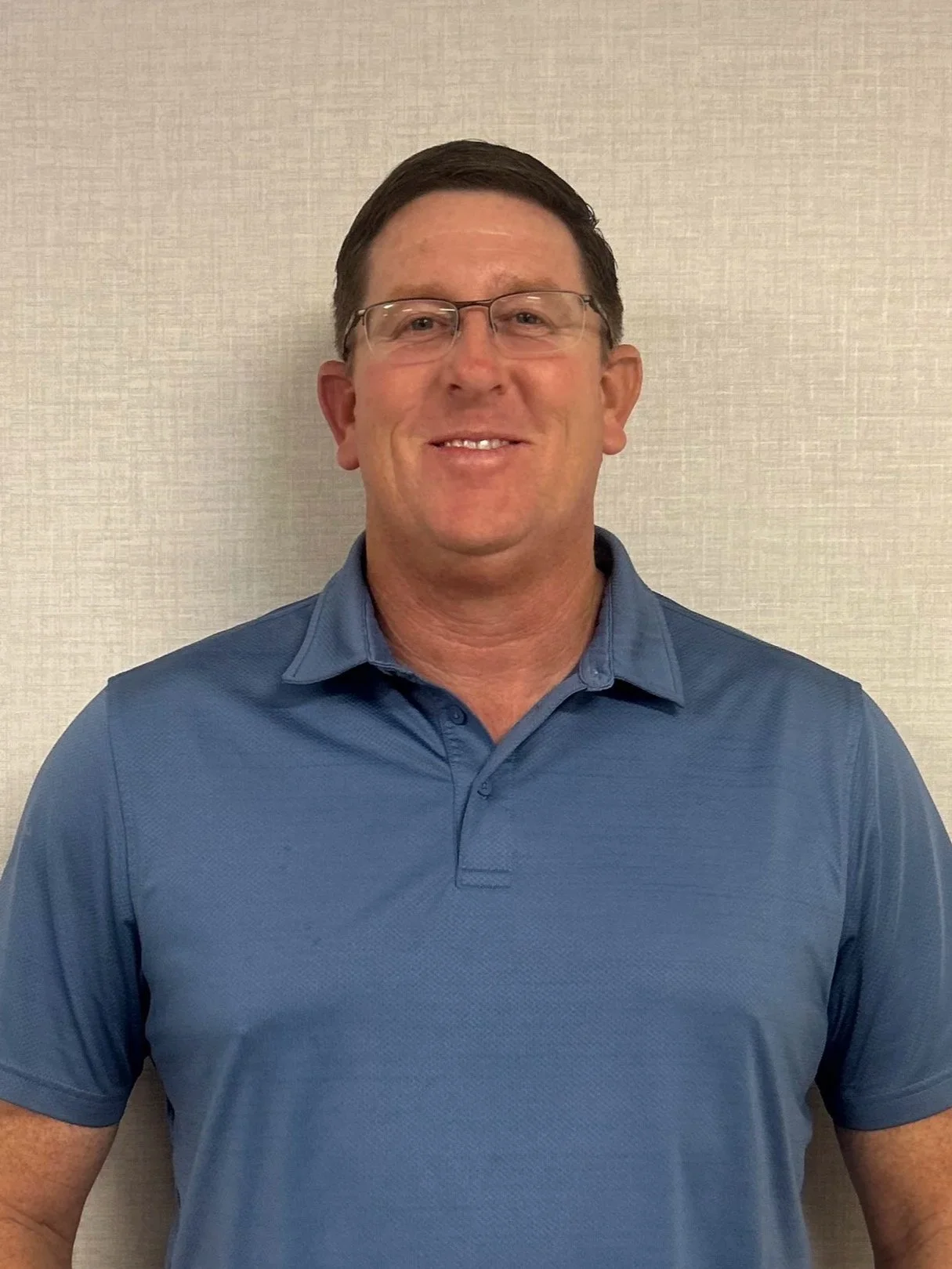 A portrait of a middle-aged man with glasses, short dark hair, wearing a blue polo shirt, standing in front of a beige wall.