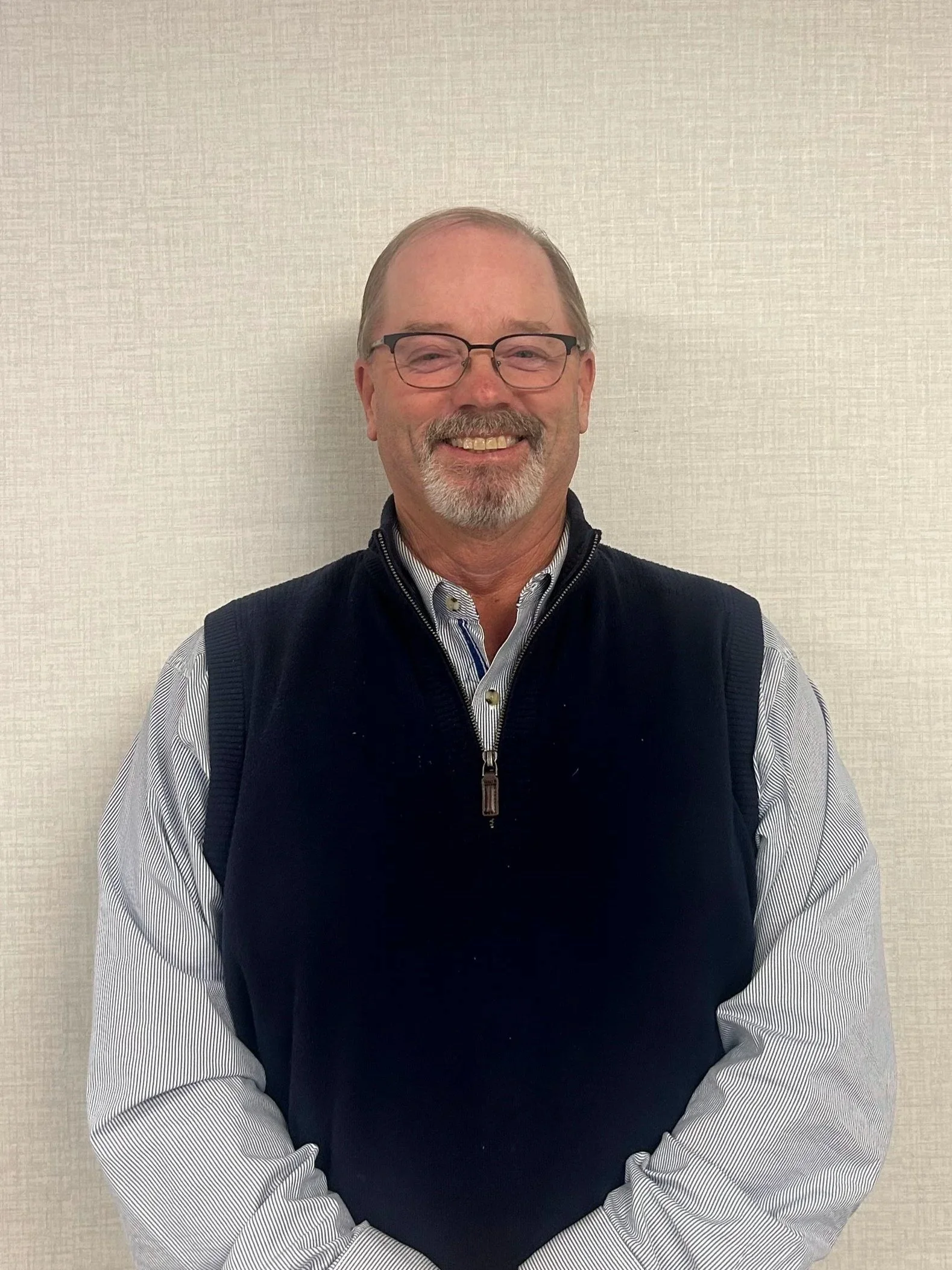 A middle-aged man with glasses, a mustache, and a beard, smiling while wearing a blue vest over a striped shirt, standing against a plain beige wall.