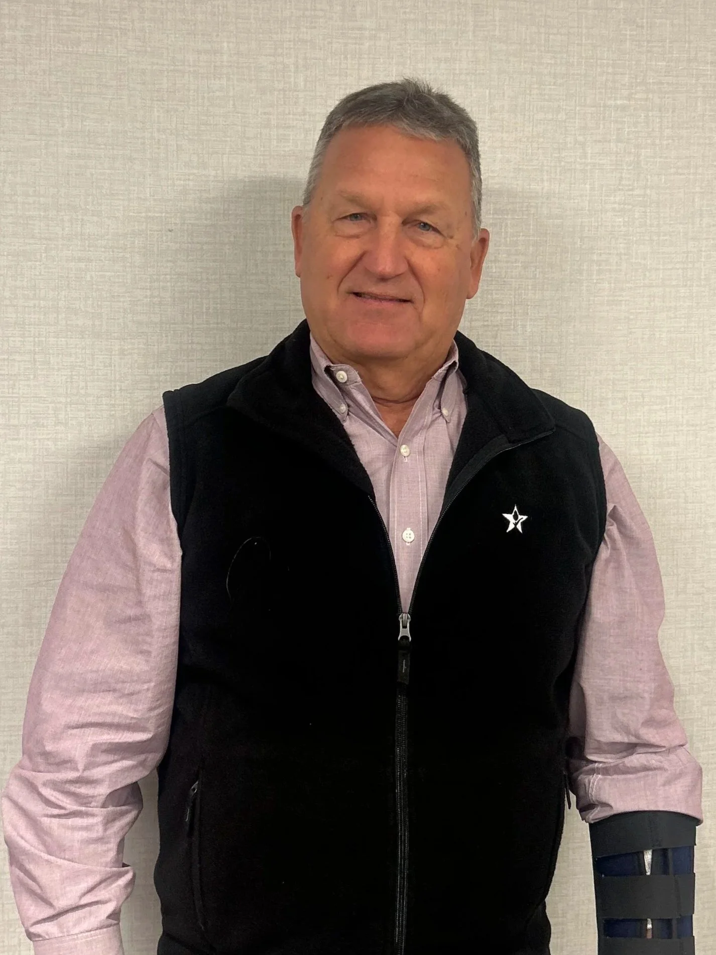 A middle-aged man with short gray hair smiling, wearing a pink button-up shirt and a black vest with a star logo, standing against a plain beige wall.