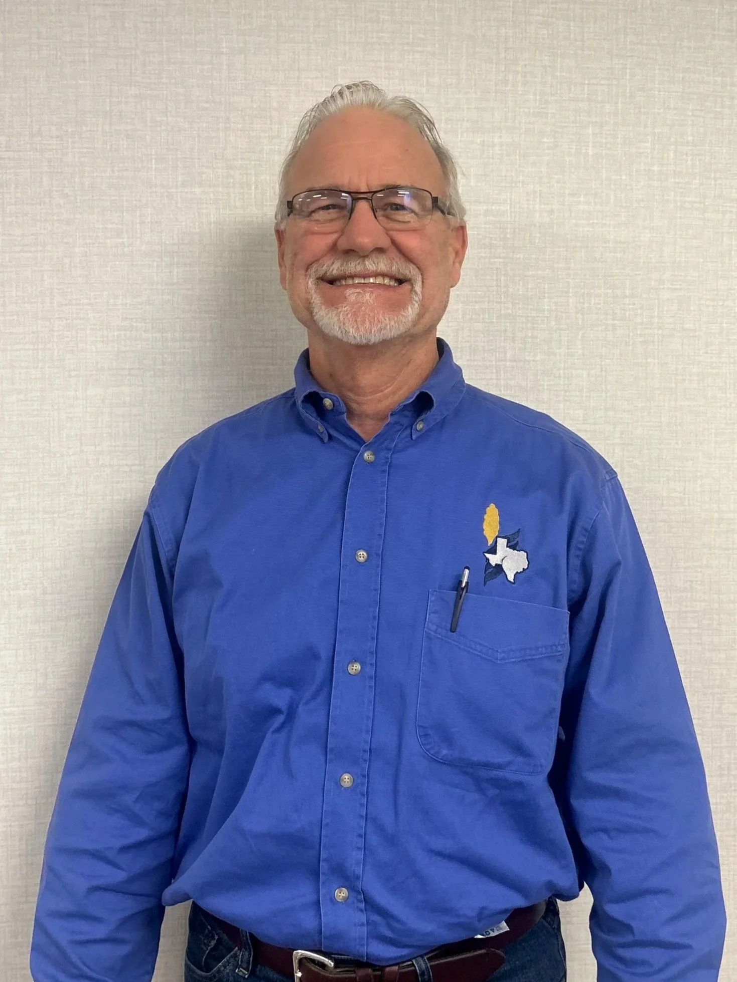 A smiling older man with gray hair, glasses, and a beard, wearing a blue button-up shirt with an Alabama and Texas state emblem on the chest, standing against a plain beige wall.