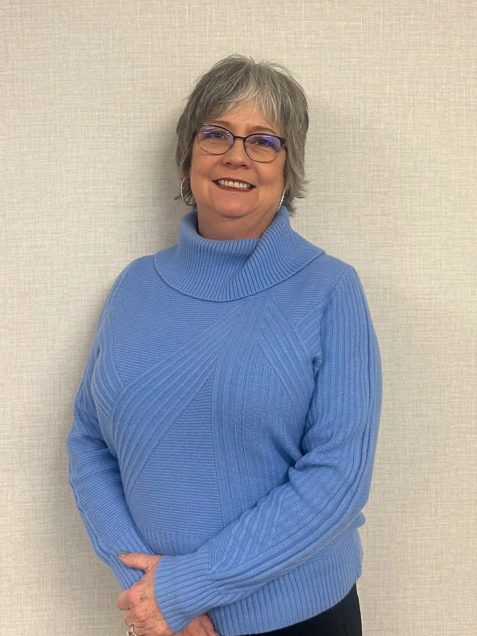A smiling middle-aged woman with short gray hair and glasses, wearing a blue turtleneck sweater, standing against a neutral wall.