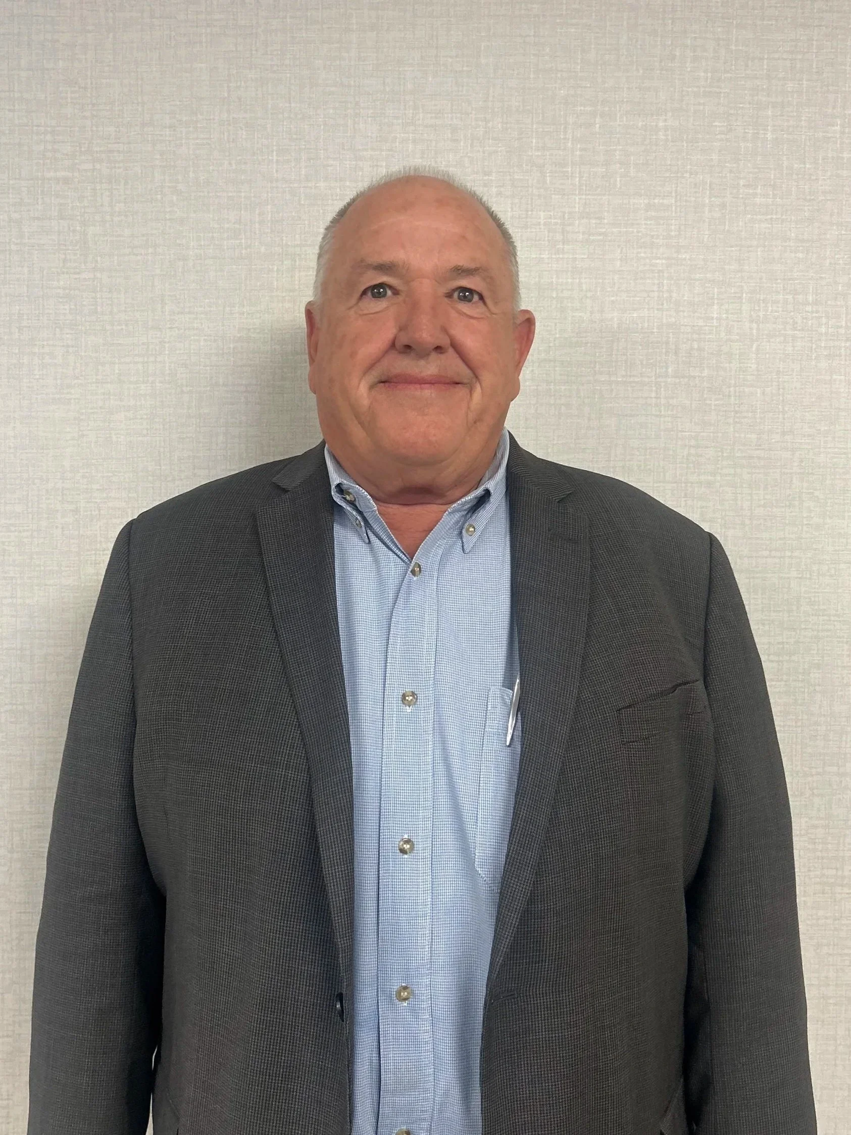 A middle-aged man wearing a dark blazer and a light blue collared shirt, smiling, stands against a plain light-colored wall.