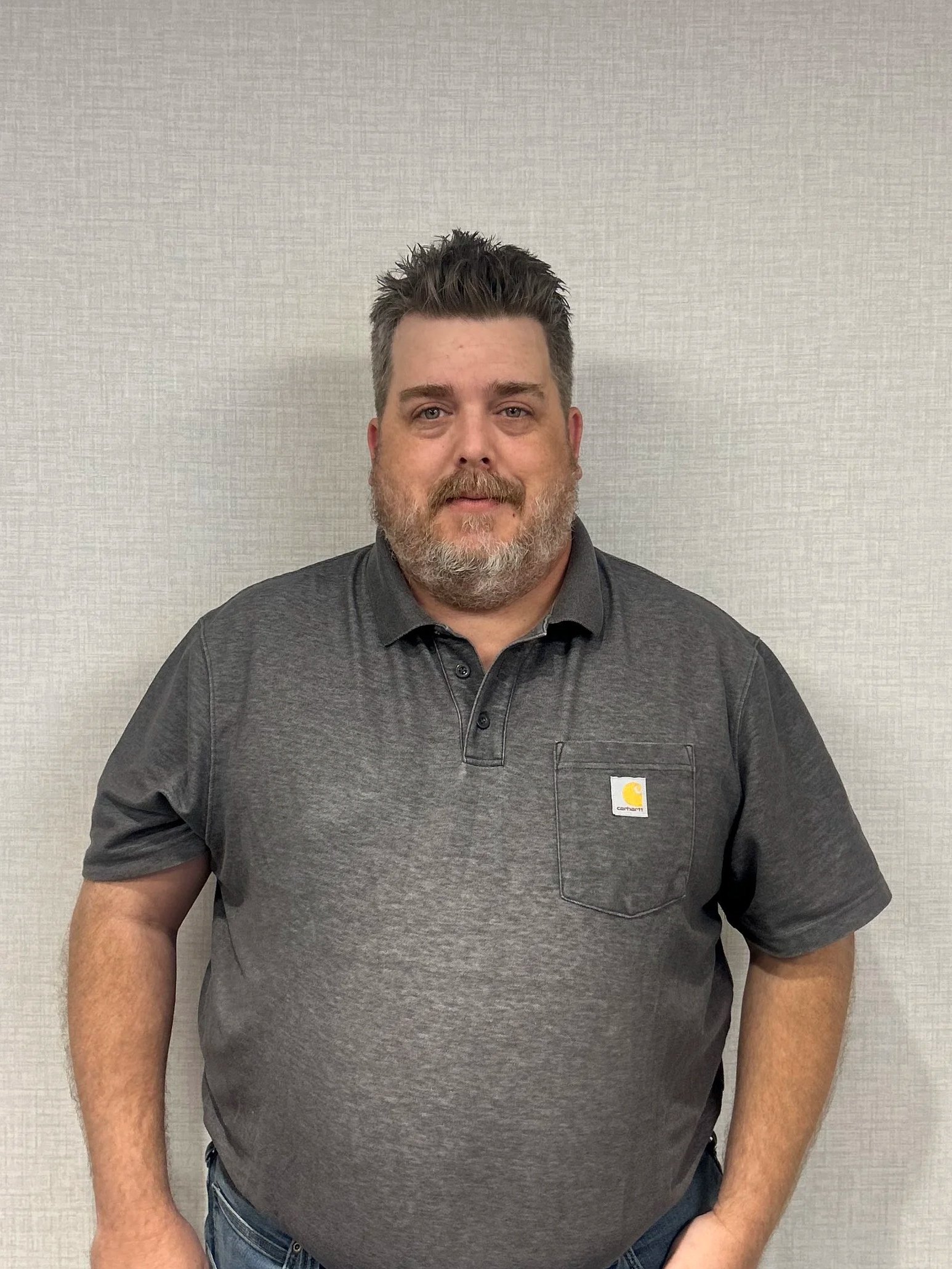 A middle-aged man with short, spiked, salt-and-pepper hair and a beard, wearing a dark gray Polo shirt with a Carhartt logo on the front pocket, standing against a plain textured wall.