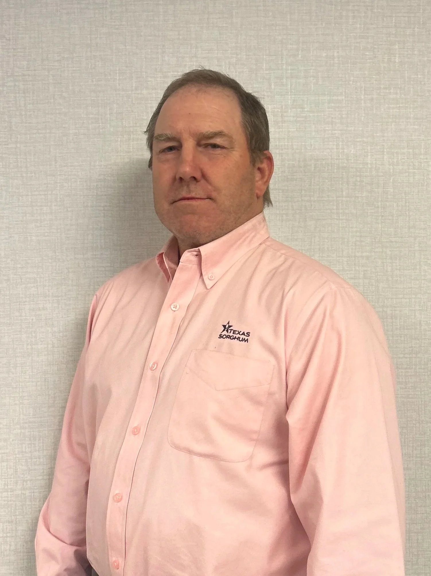 A man in a pink button-up shirt with a Texas Sorghum logo, standing against a plain light-colored wall.