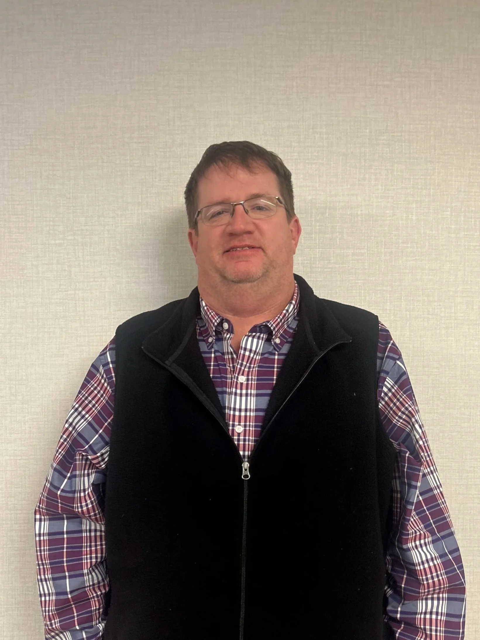 A man wearing glasses, a checkered shirt, and a black vest standing against a plain light-colored wall.