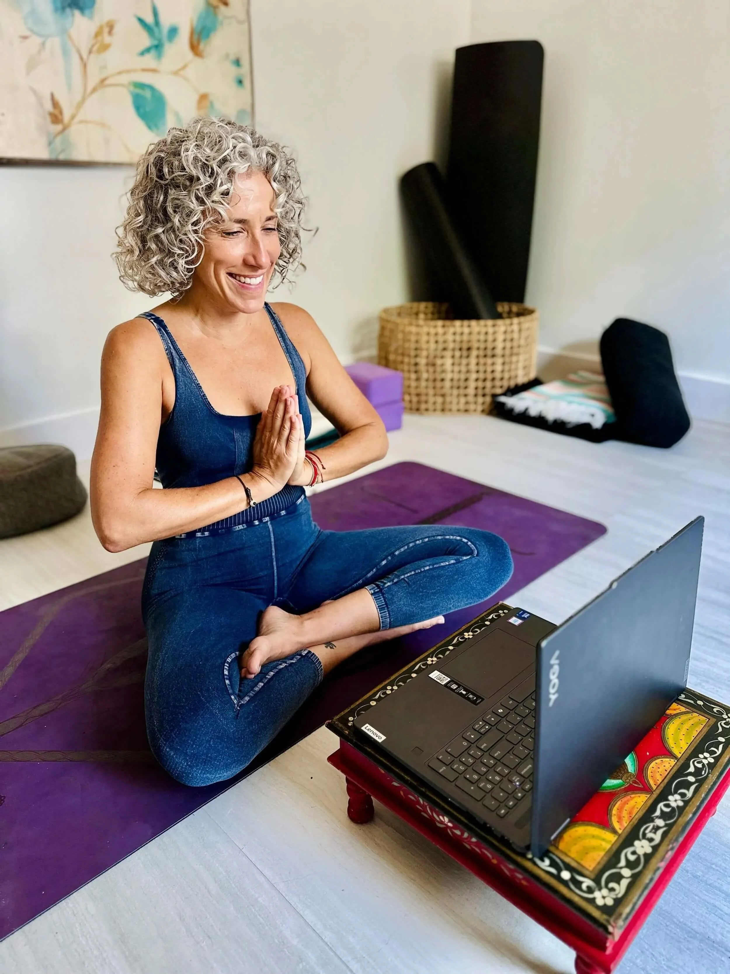Jacqueline Burbage, a somatic and yoga therapist, practicing yoga indoors, sitting cross-legged on a purple yoga mat, with her hands in prayer position while looking at a laptop in front of her.
