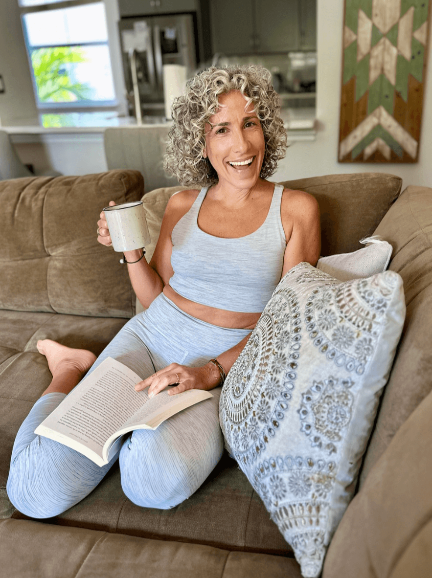 Jacqueline Burbage, a somatic and yoga therapist, smiling and holding a mug while sitting on a couch, with an open book on her lap in a cozy living room.