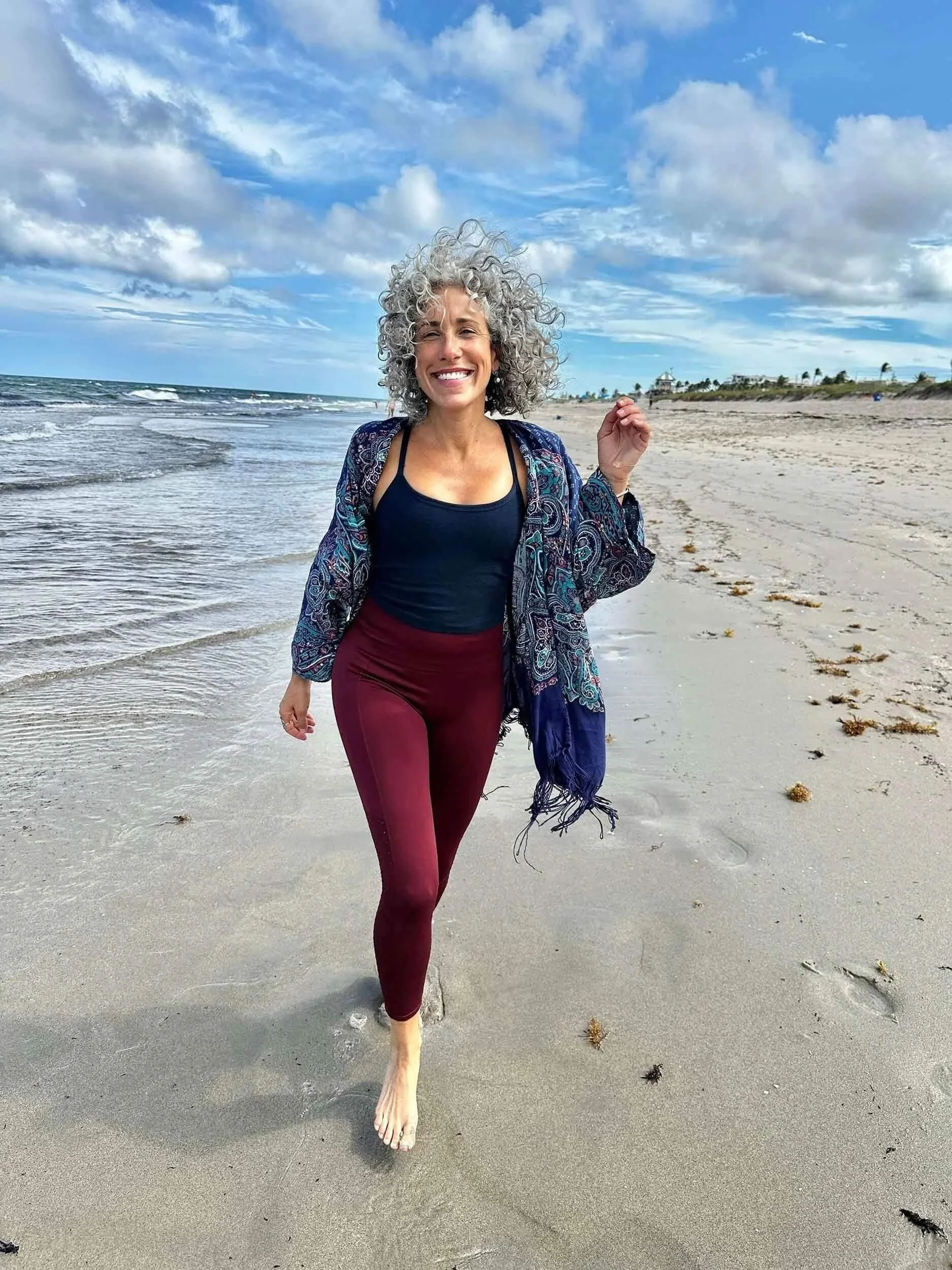 Jacqueline Burbage, a somatic and yoga therapist, walking barefoot on the beach, smiling, with a blue sky and clouds overhead, and the ocean in the background.
