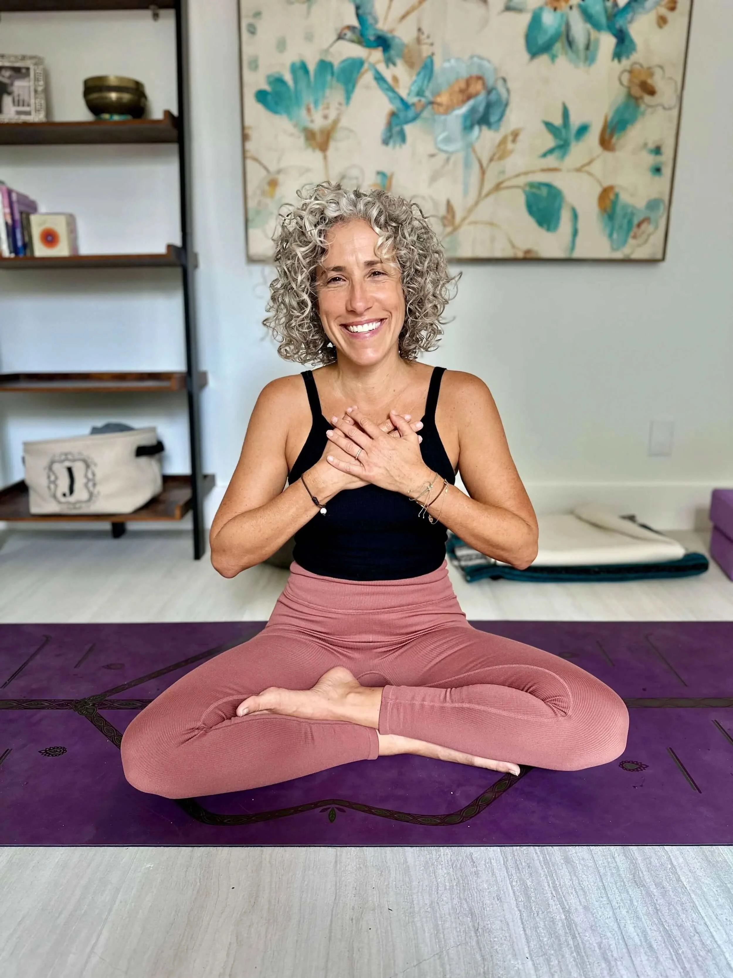 Jacqueline Burbage, a somatic and yoga therapist, sitting on a purple yoga mat in a cross-legged yoga pose, smiling with her hands on her chest.