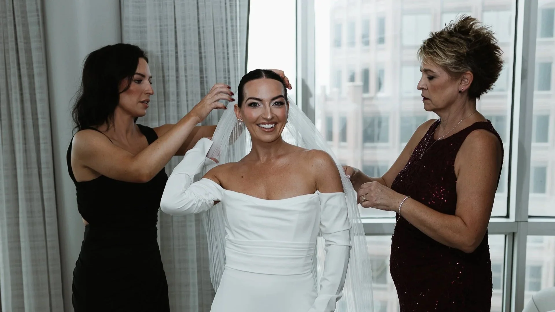 A bride in a white wedding dress and veil is being dressed by two women in a room with large windows behind them.