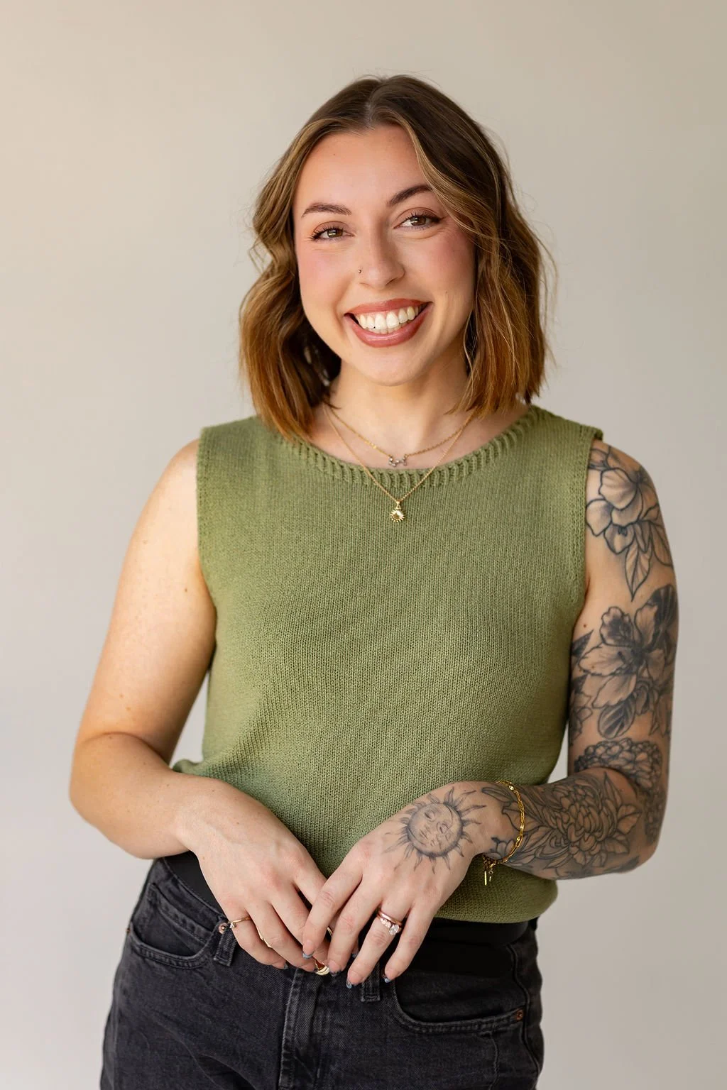 A smiling woman with shoulder-length brown hair, wearing a green sleeveless top and black jeans, showcases tattoos on her right arm, and accessorizes with layered necklaces and bracelets.
