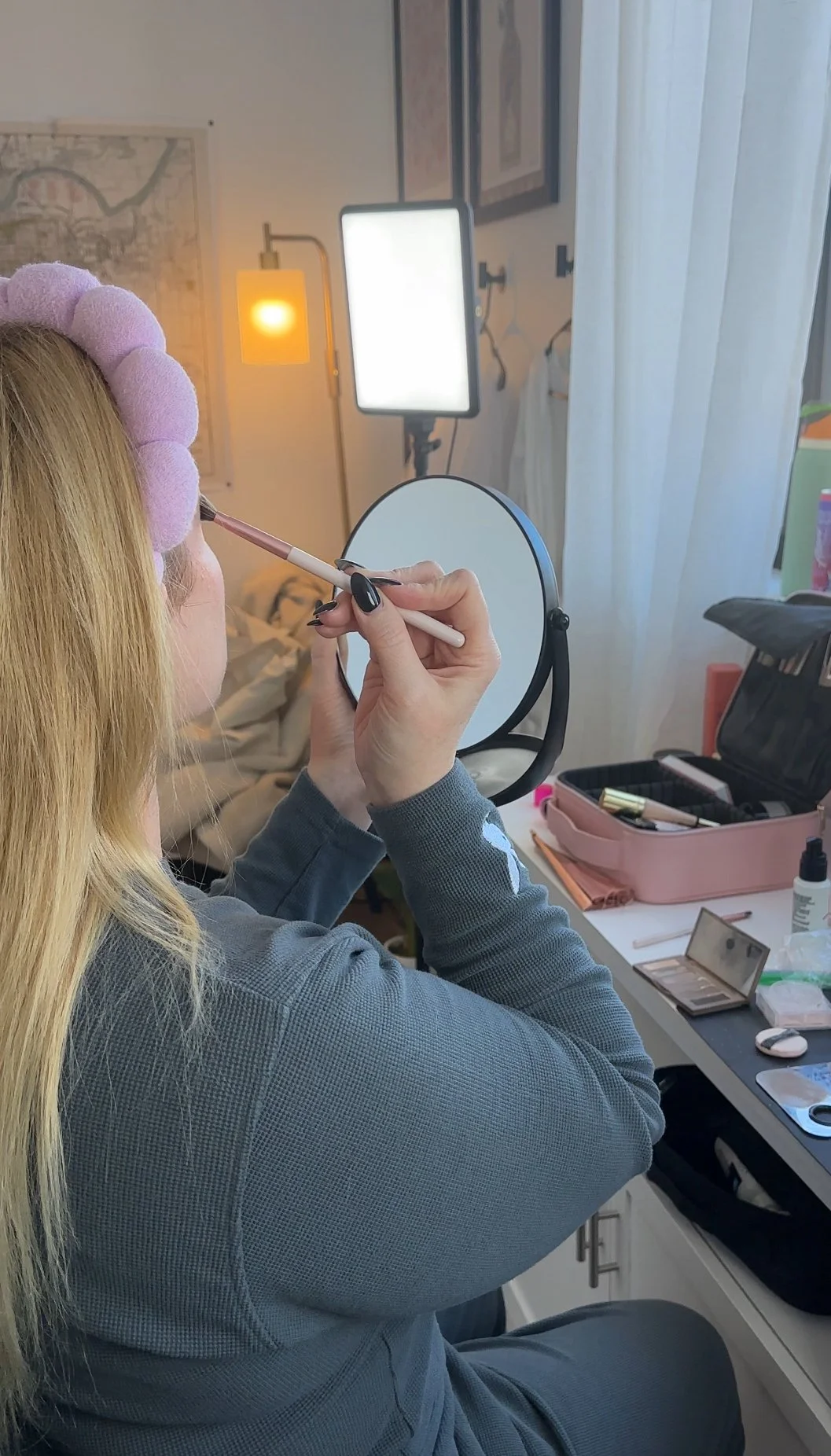 A woman sitting at a makeup station with a ring light, applying makeup while looking into a mirror.