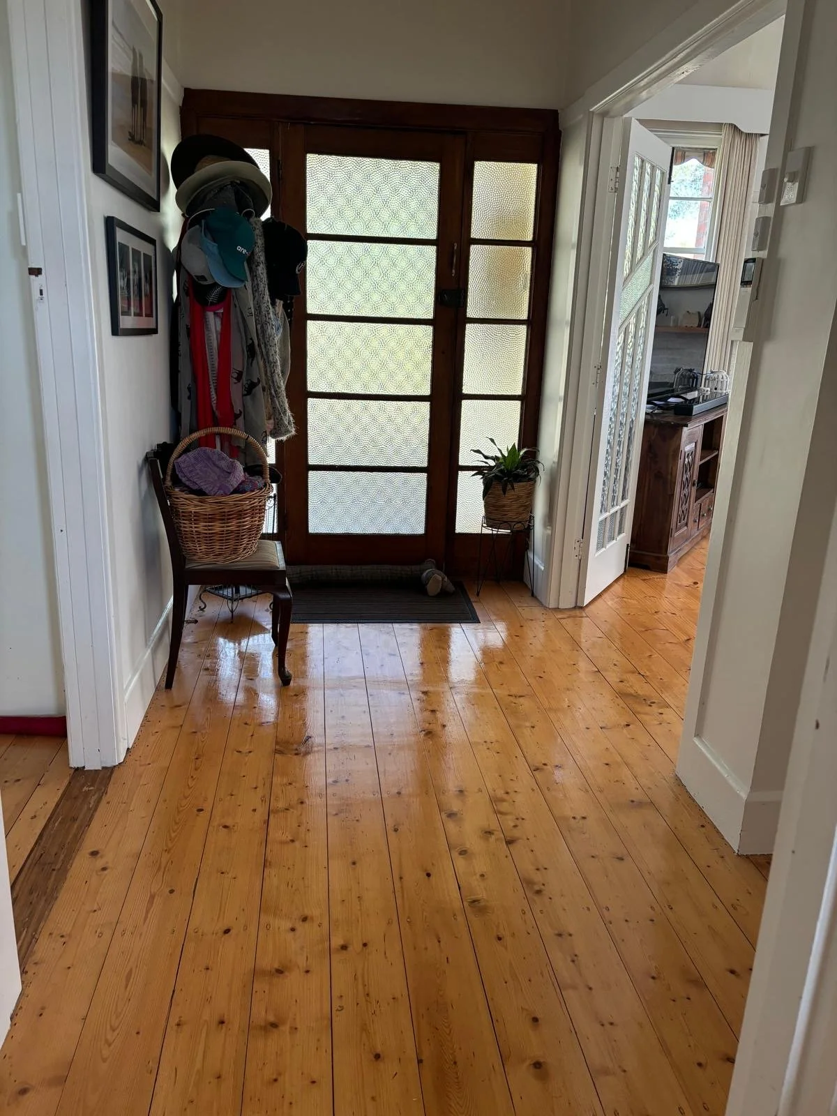 Entrance hallway with wooden floor and door with frosted glass panels, coat rack with hats, scarves, and umbrellas, a small chair with a basket on top, a potted plant near the door, and a glimpse into another room with a wooden sideboard.