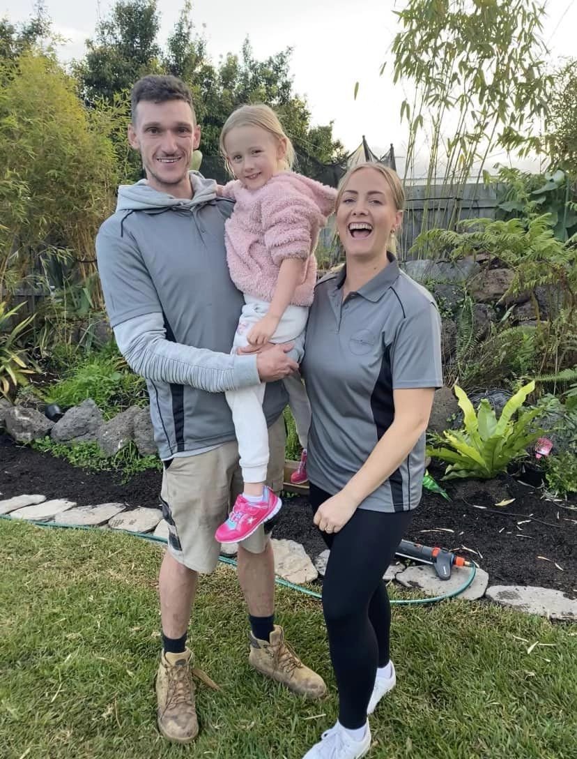 A family of three standing outdoors in a garden, smiling. The man is holding a young girl, and a woman stands next to them.