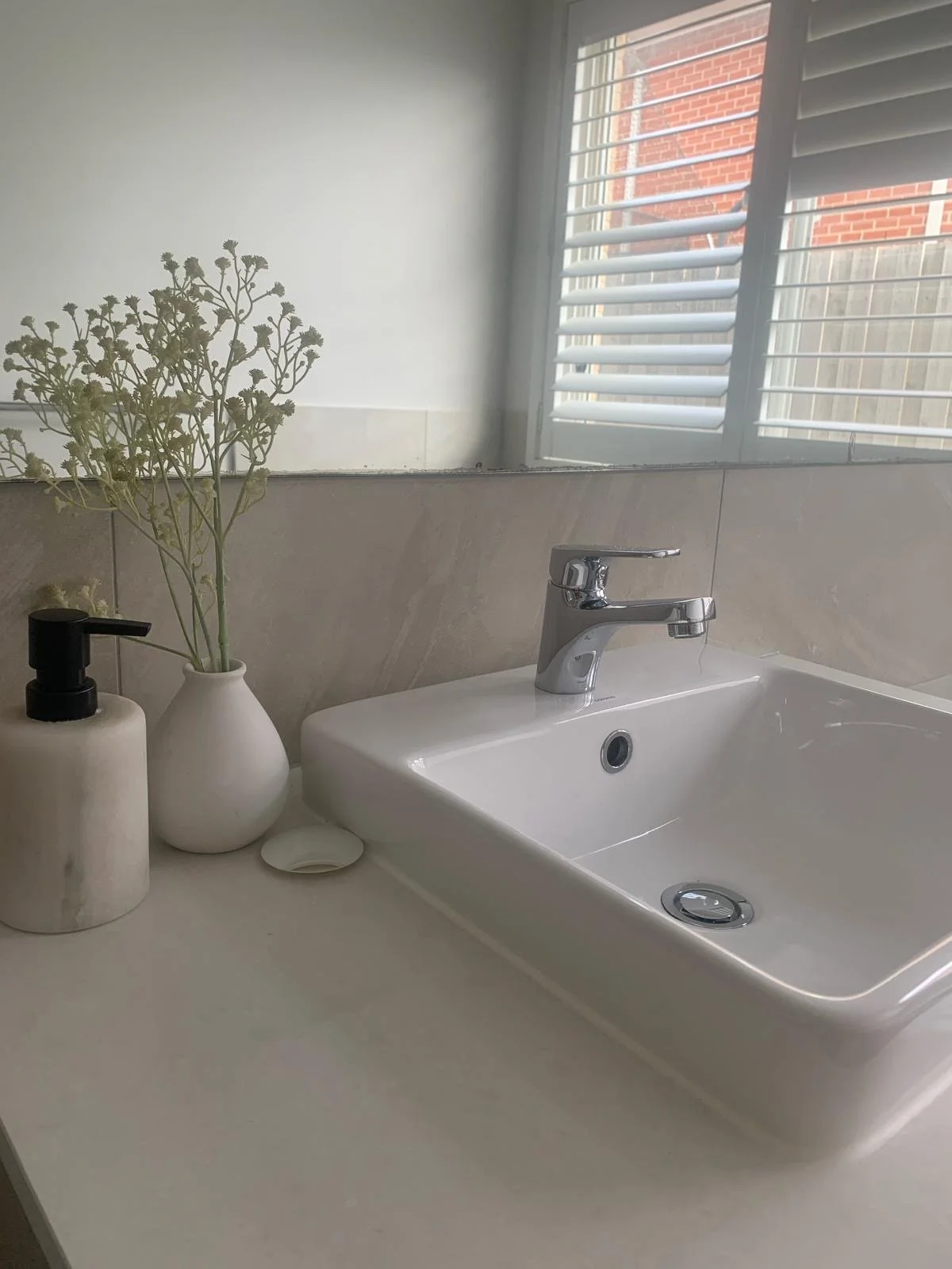 A bathroom countertop with a white rectangular sink, a chrome faucet, a black soap dispenser, a small white dish, a white vase with green foliage, and a mirror reflecting a window with white shutters and a brick wall outside.