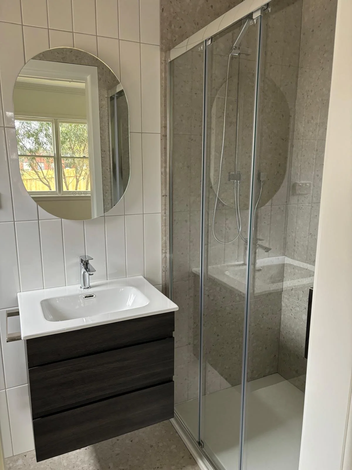 A modern bathroom with a white sink on a dark wood vanity, oval mirror, and a glass shower enclosure with a showerhead and a window reflecting outside greenery.