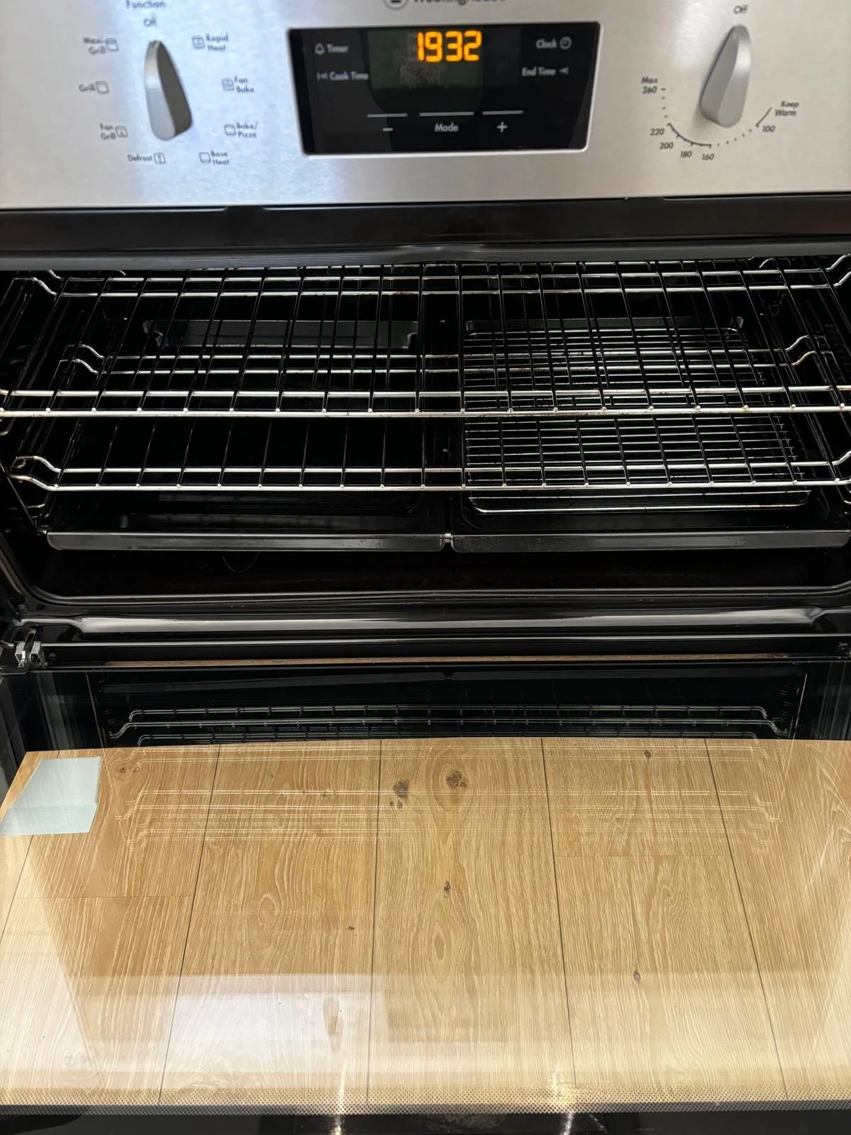 An empty modern oven with a digital clock displaying 19:32, digital and rotary controls on the top panel, and a wooden cutting board on the oven door.
