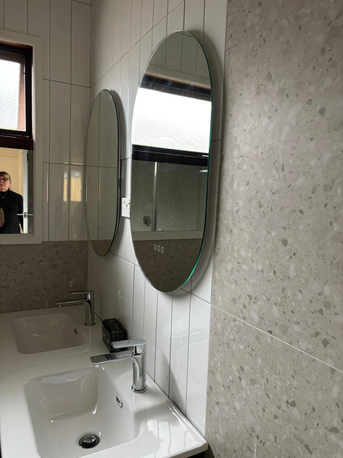 A bathroom with two oval mirrors above a white double sink vanity with silver faucets, a beige tiled wall, a small window, and a person taking a photo reflected in the mirror.