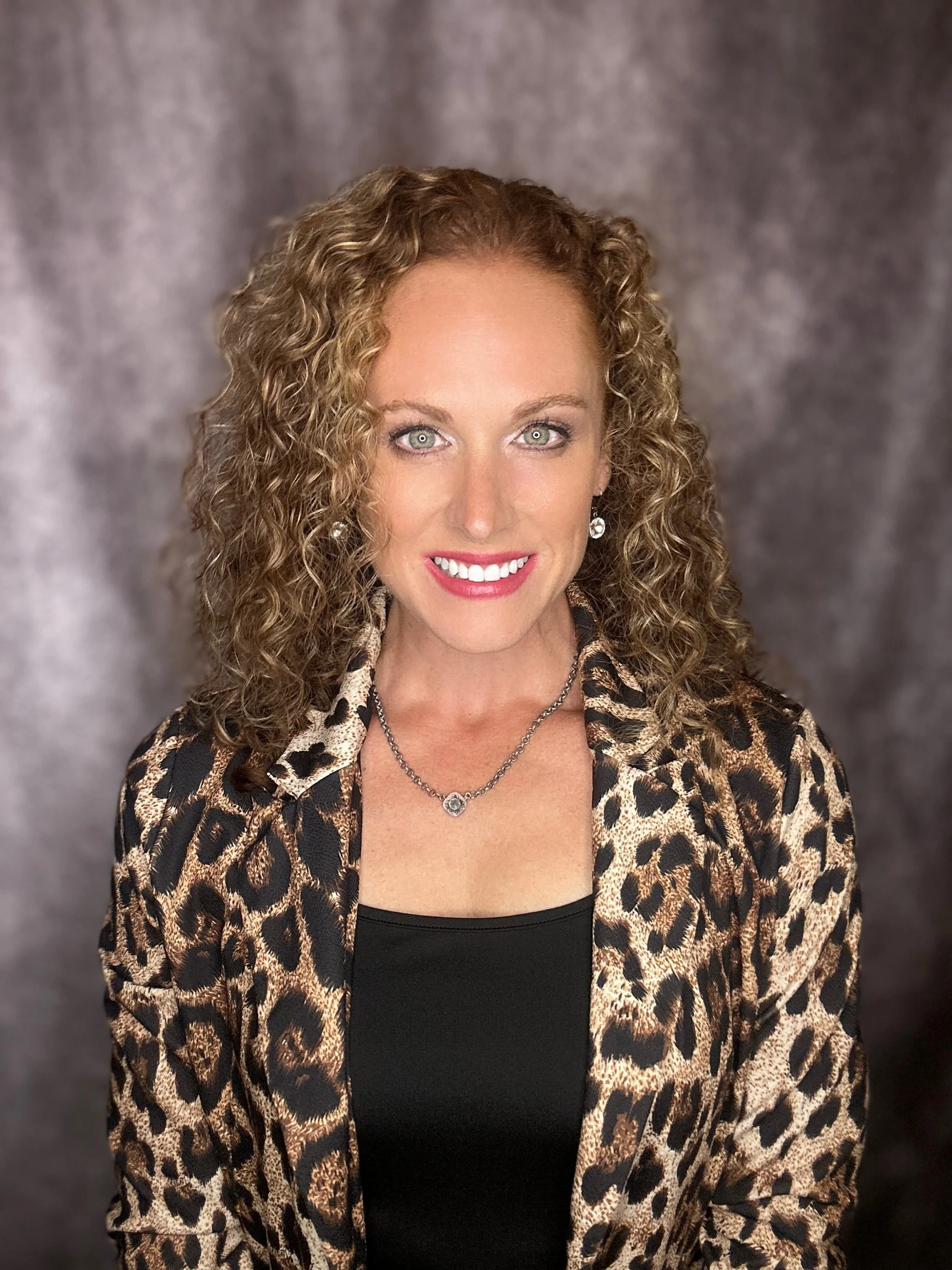 Woman with curly hair wearing a leopard print jacket and black top, smiling against a dark background.