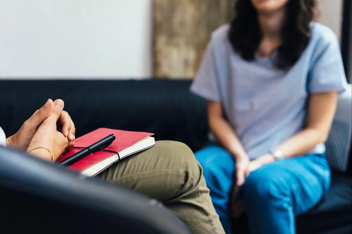 Two people sitting in a therapy session, one holding a red notebook with a pen, hands clasped. The other person is partially visible, sitting across.
