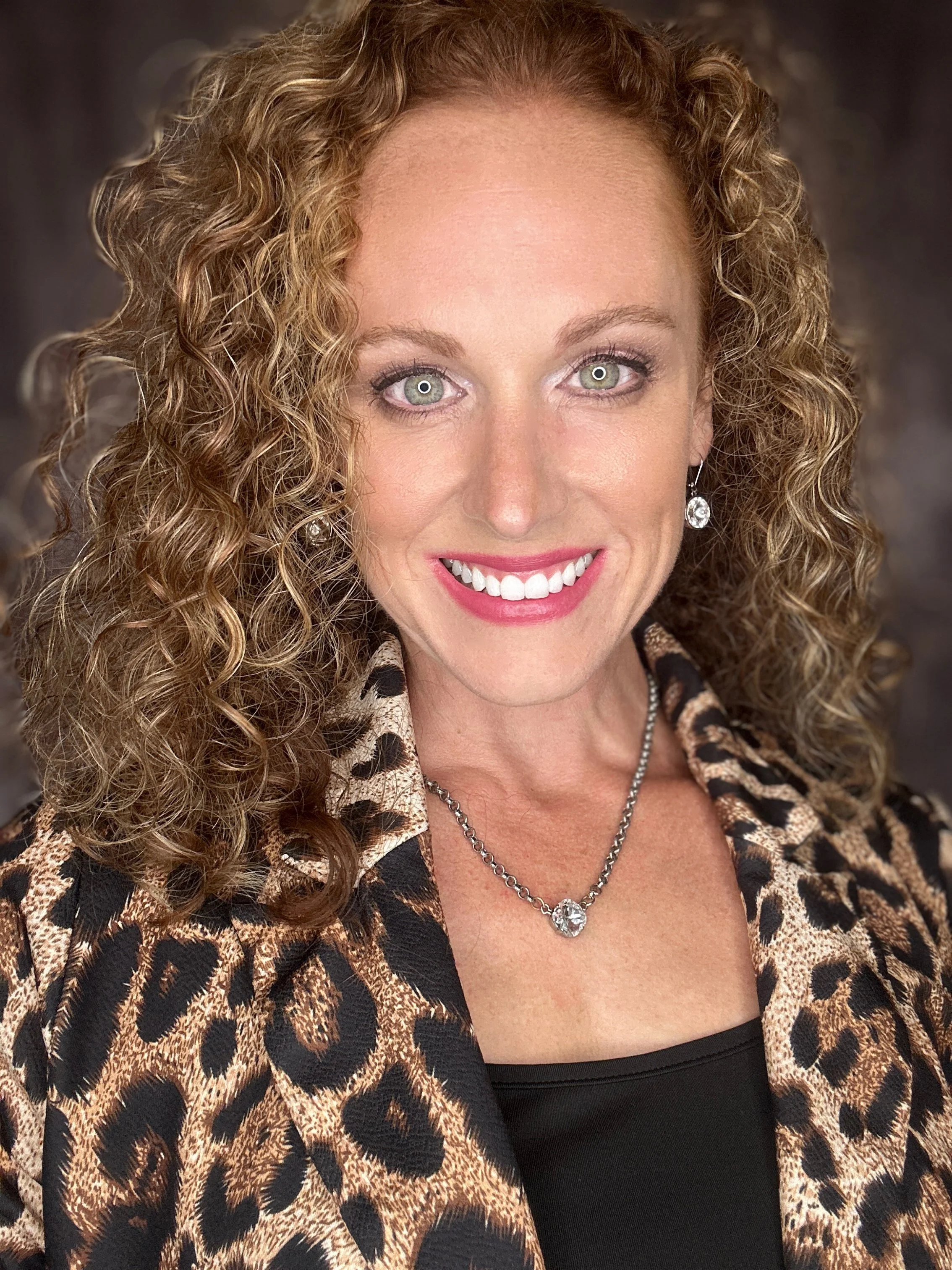 Smiling woman with curly hair wearing a leopard print jacket and a necklace.