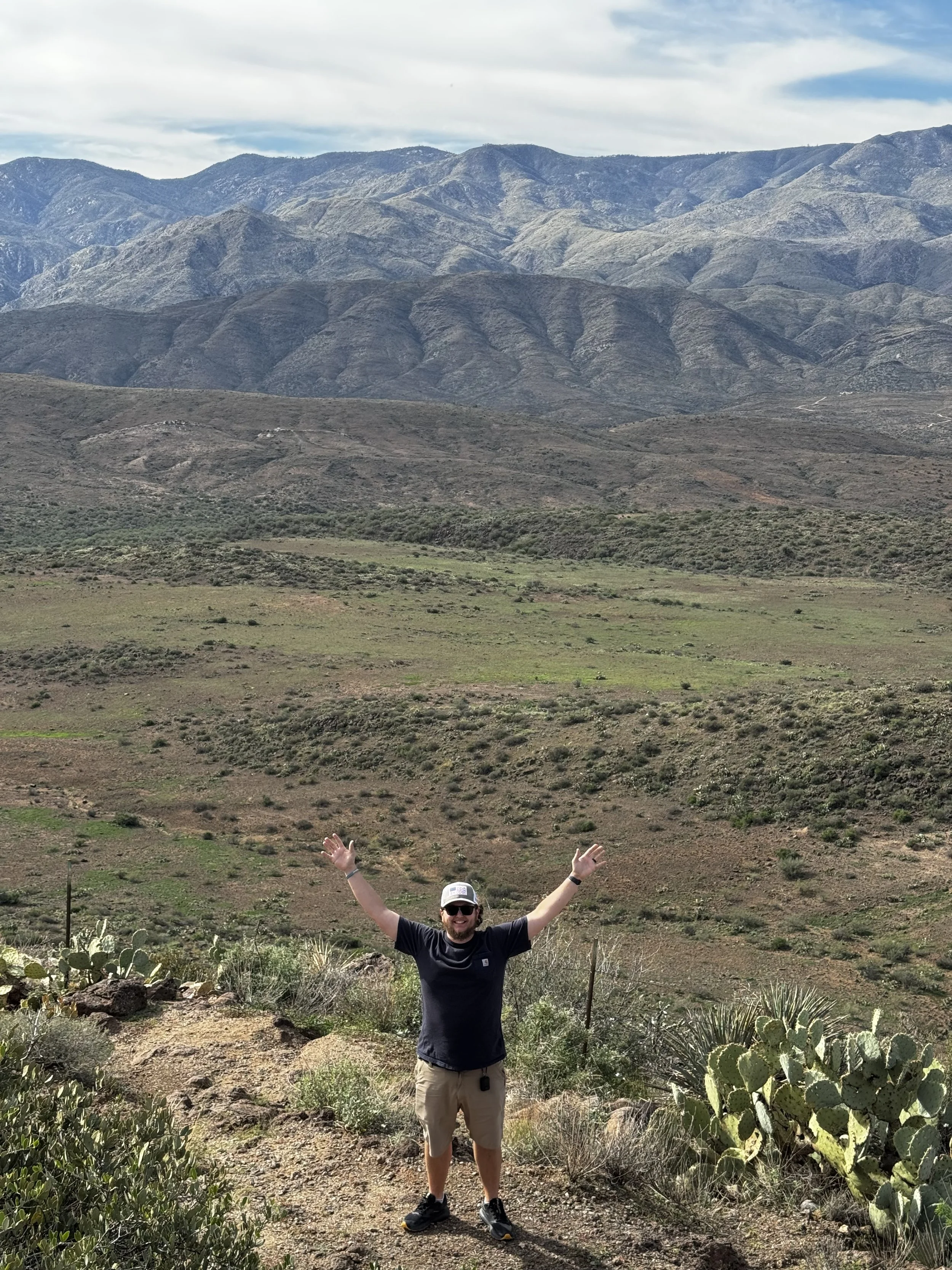 A man wearing a black shirt, khaki shorts, and a hat stands in a desert landscape with his arms raised, surrounded by cacti and desert plants, with mountains and a partly cloudy sky in the background.