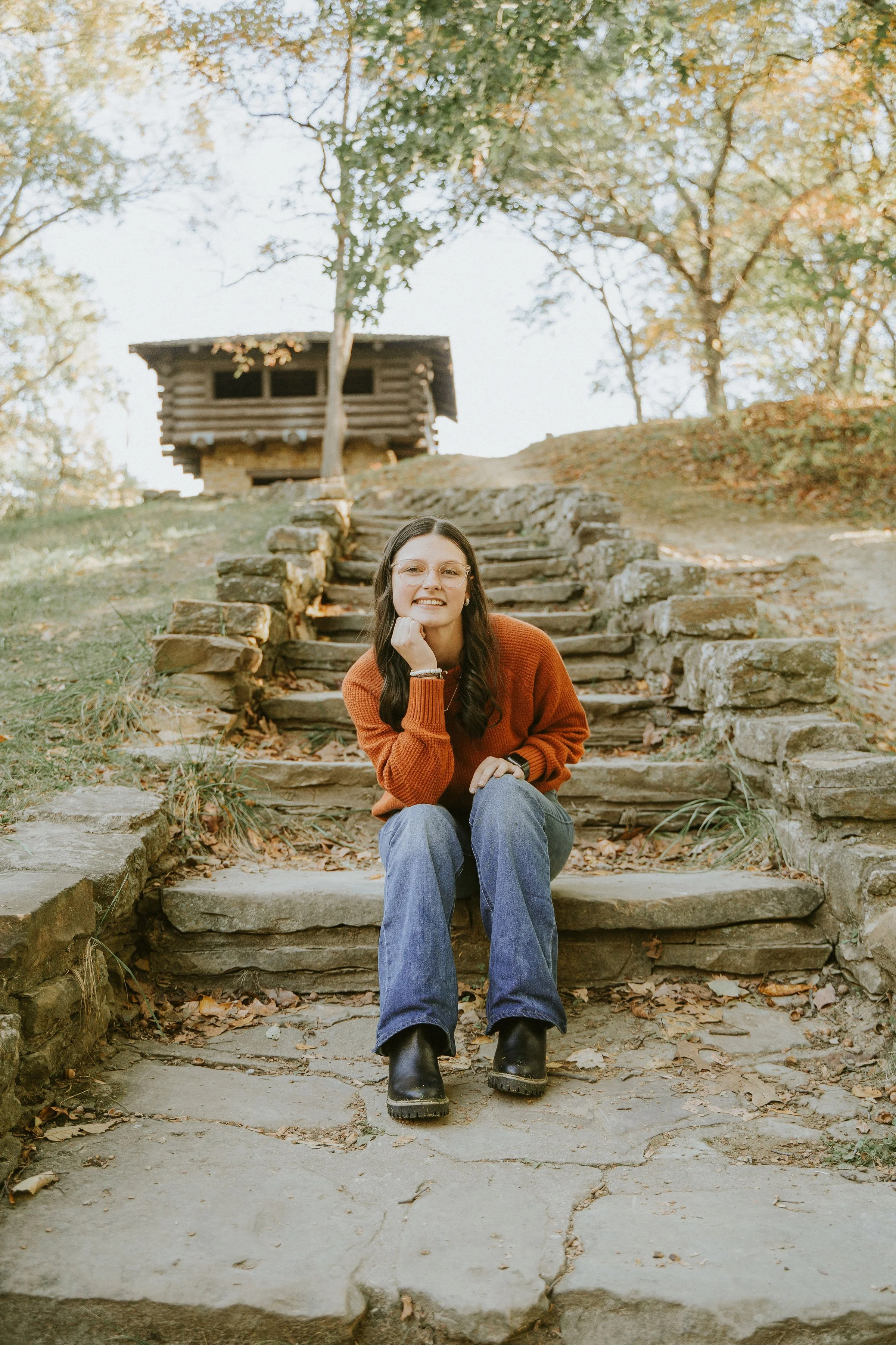 A woman with long dark hair wearing glasses, an orange sweater, and blue jeans, sitting on stone steps outdoors in autumn, smiling at the camera.