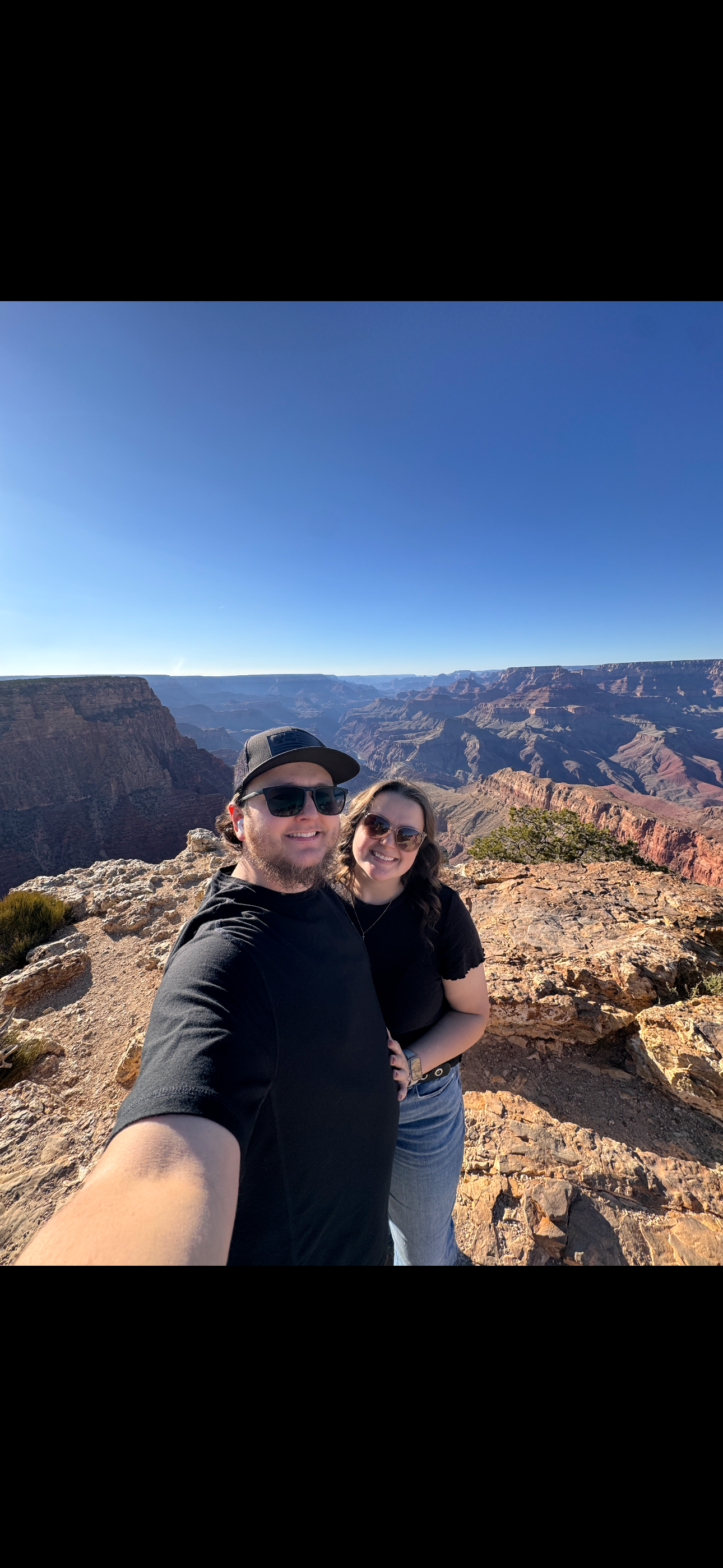 Two people taking a selfie at the Grand Canyon with the canyon's layered rocks and deep valleys visible in the background under a clear blue sky.