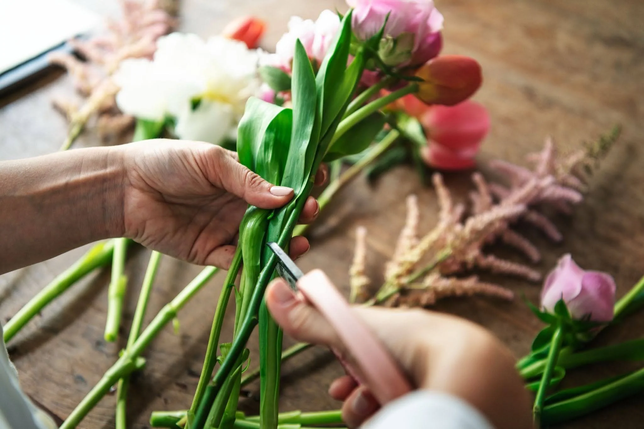 Hands trimming fresh pink and white flowers with scissors on a wooden table, preparing a floral bouquet