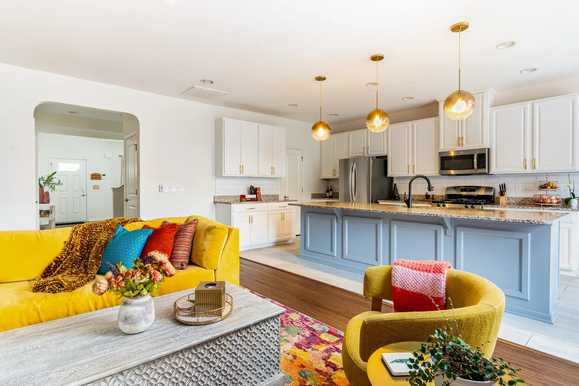Open-concept living room and kitchen with white cabinets, stainless steel appliances, pendant lights, and colorful furniture including yellow couches and an orange chair.