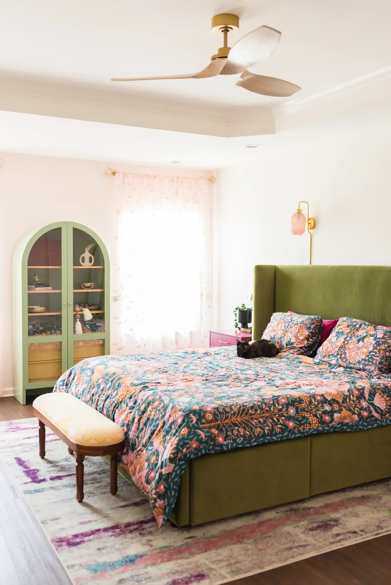 A bedroom featuring a green upholstered bed with a colorful floral bedspread, a small yellow and wood bench at the foot of the bed, a pink nightstand with a black plant pot, a pink wall sconce, a green arched cabinet with glass doors, and a window with sheer pink curtains,  a black cat lying on the bed, and a patterned area rug.
