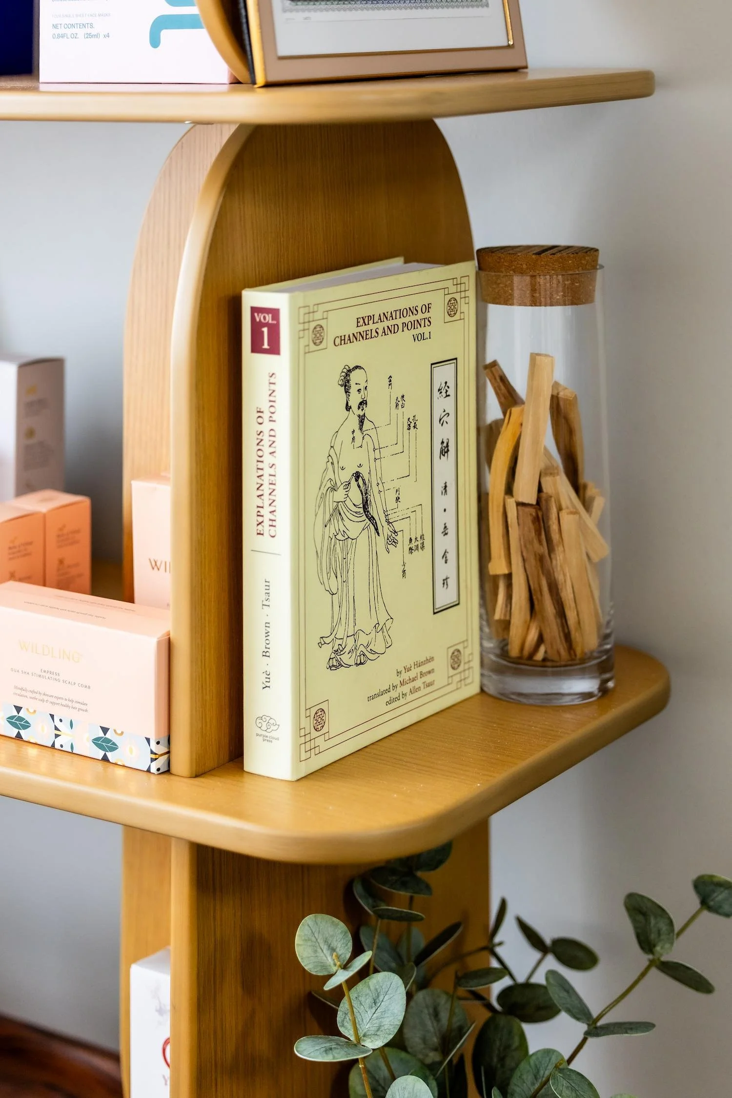 Wooden corner shelf holding books, a glass jar with wooden sticks, and a light pink product box with a patterned bottom edge, with eucalyptus leaves in the foreground.