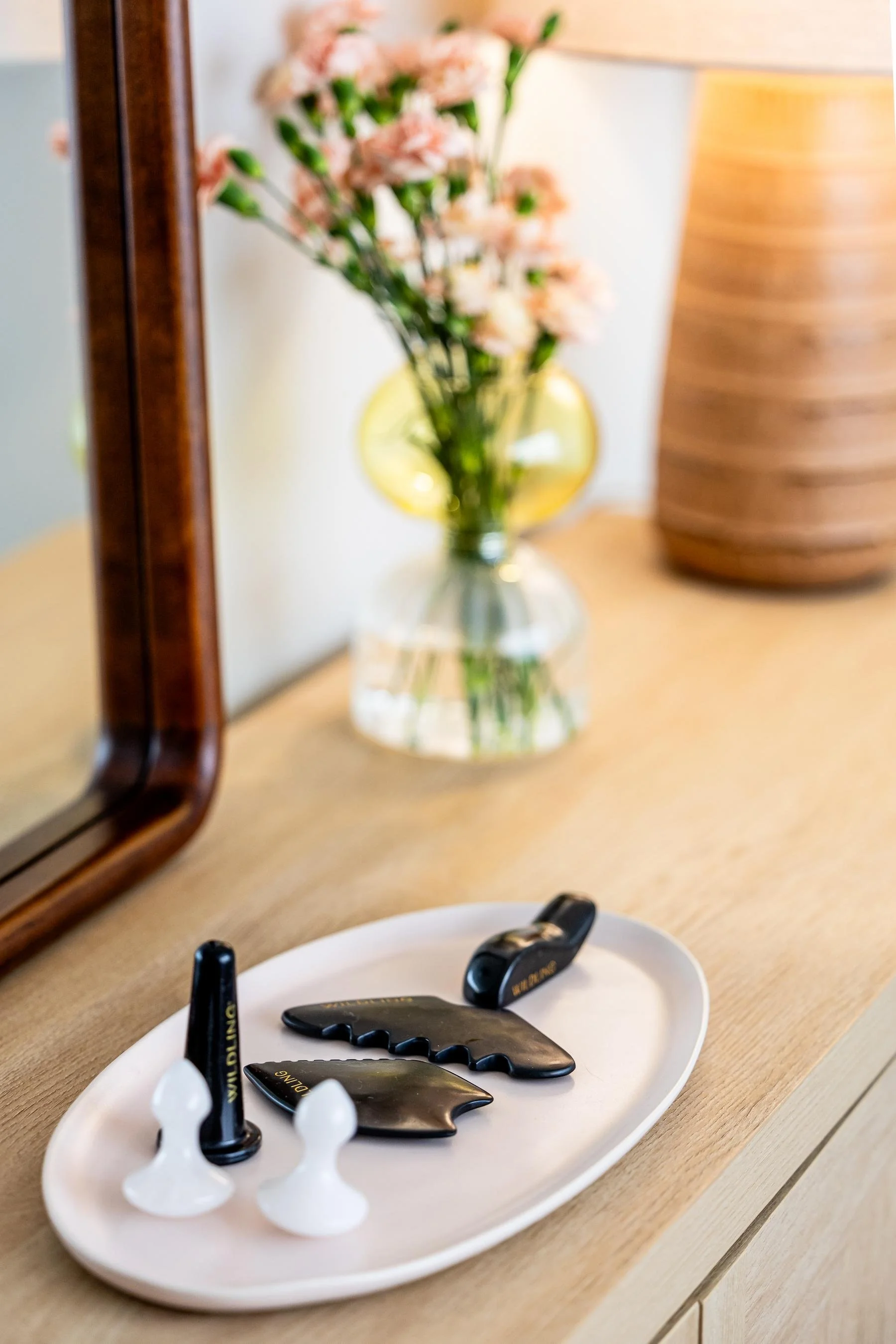 A white plate with chocolate syrup cookies and black edible writing pens, placed on a wooden surface, with a mirror, a glass vase with pink flowers, and a lampshade in the background.