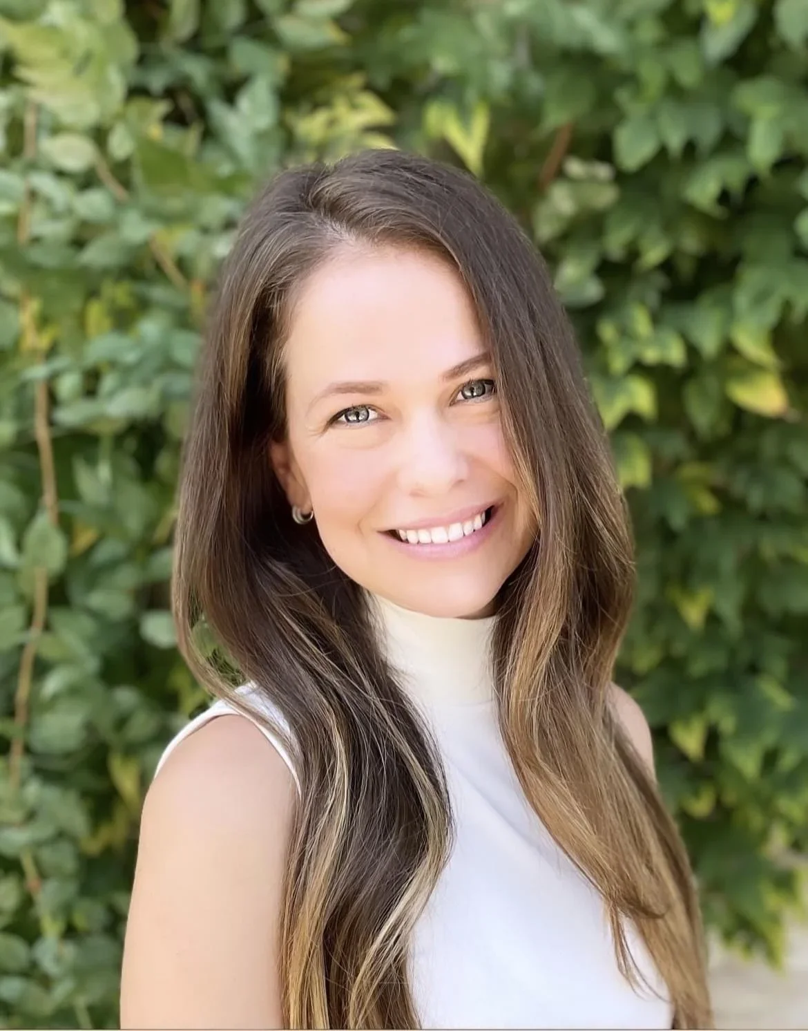An interior design client with long brown hair, wearing a sleeveless white top, standing outdoors with green foliage in the background.