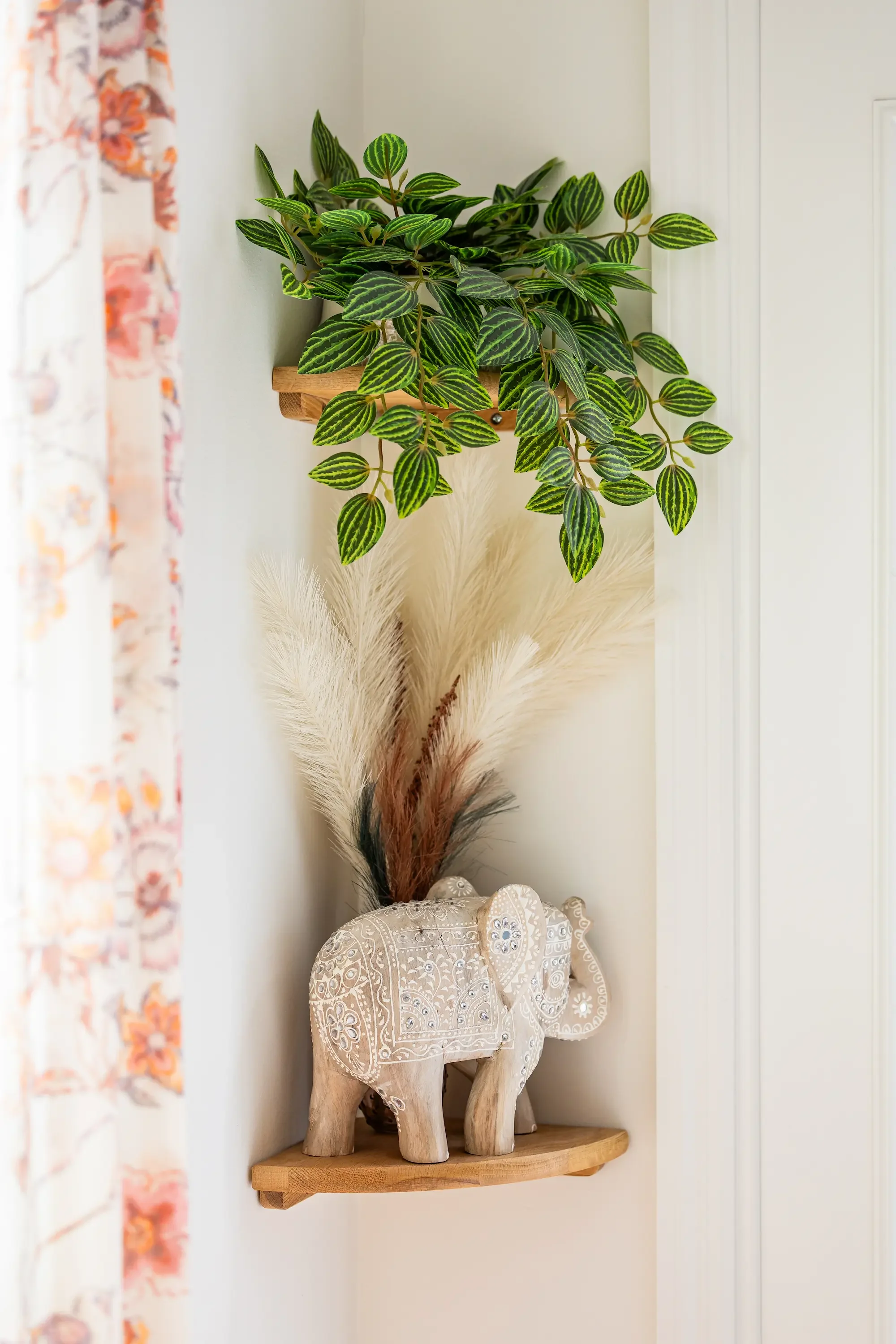 A decorative wooden elephant with intricate white embroidery patterns standing on a wooden shelf. Behind it, there is a bouquet of tall, lush green leaves with striped patterns, placed in a wall-mounted wooden planter. The shelf and plant are against light-colored walls, with a floral patterned curtain partially visible on the left side of the image.